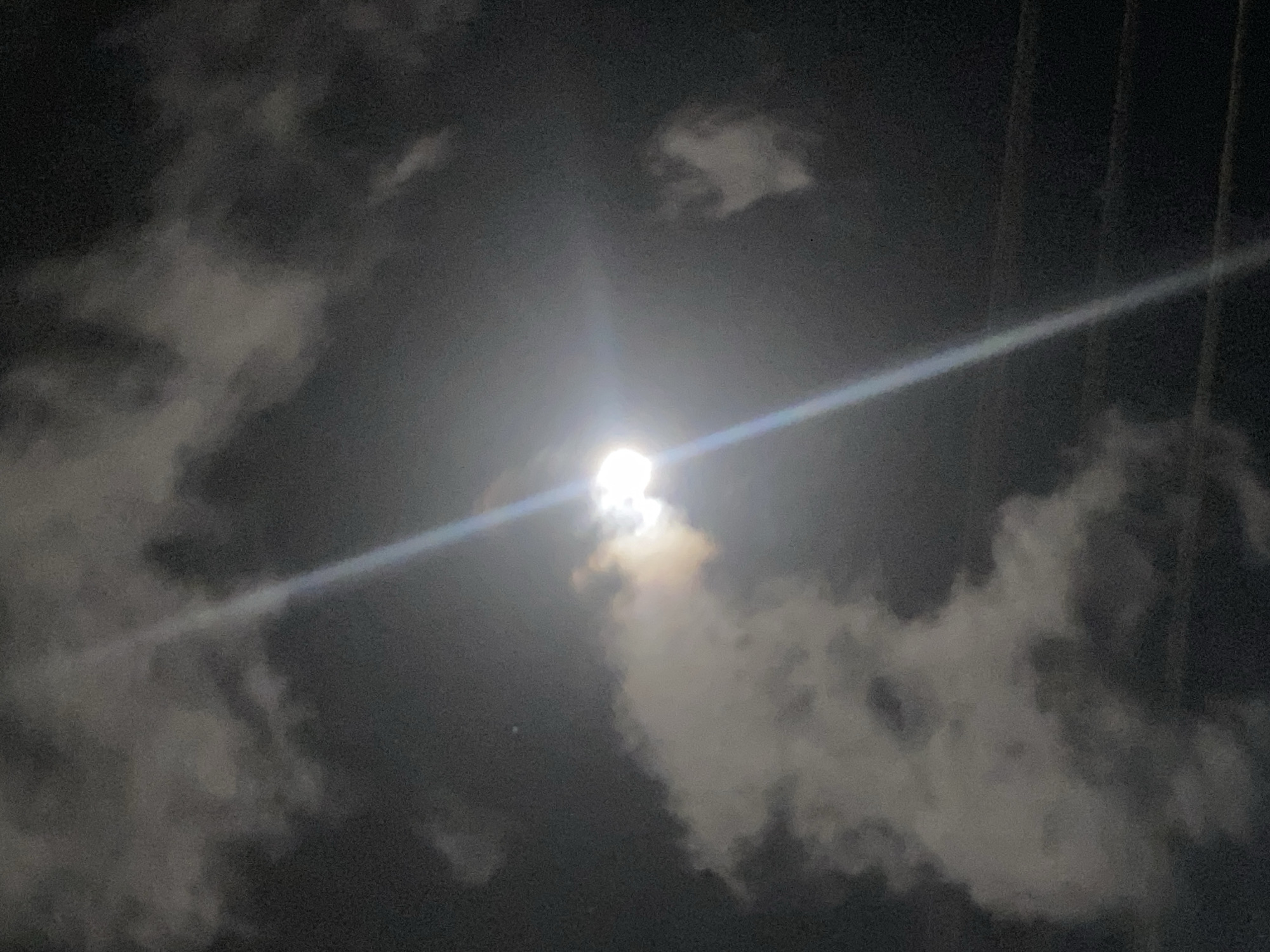 The image depicts a night scene in Juan Griego, Venezuela, featuring a bright moon partially obscured by clouds. Two prominent linear features, likely power lines, diagonally cross the upper portion of the frame. The sky is dark with varying cloud formations, some illuminated by the moon.