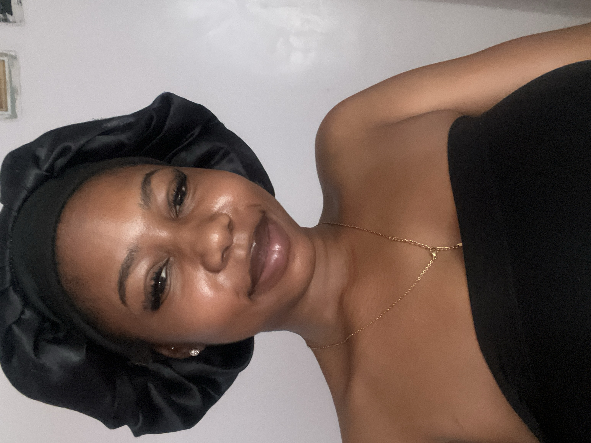 A medium shot shows a young Black woman with dark skin, wearing a black satin bonnet and a black strapless top. She has on a thin gold chain with a small pendant. Her makeup includes defined eyebrows, eyeliner, and lip gloss. A small diamond stud earring is visible on her left ear. The background is a plain white wall, with some indistinct framed objects visible at the top. The lighting is soft and even. No information about the location in the city of Rafin Par, Nigeria, is available.
