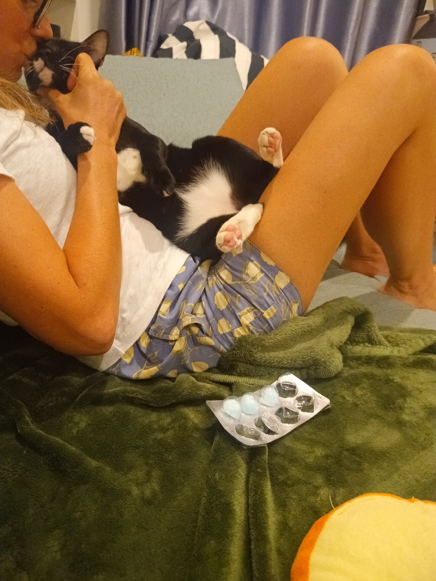 This indoor image, likely taken in Ko Pha-ngan, Thailand, captures a tender moment of affection between a person and their cat. A person, appearing female with blonde hair and wearing glasses, a white t-shirt, and blue patterned shorts, is reclining comfortably. She is gently holding and nuzzling a black and white tuxedo cat, whose head is close to her face, suggesting a kiss or affectionate rub. The cat is relaxed, lying on its back on her lap and torso, with its soft pink paw pads clearly visible.
The scene is set in what appears to be a cozy living room or bedroom. A dark green, fluffy blanket or throw covers part of the person's lower body and the foreground. On this blanket, an open blister pack containing several light blue or white gel capsules is visible; some compartments are empty, indicating consumption. To the bottom right, a partial view of a soft, yellow, and orange object, possibly a cushion or toy, can be seen.
In the background, a light blue surface, likely a cushion or part of a sofa, and blue curtains contribute to the indoor setting. A striped cushion or pillow is also visible behind the person. The overall atmosphere is one of comfort, relaxation, and mutual affection between the pet owner and their cat. The lighting is soft, enhancing the peaceful mood. No visible text is present in the image.
