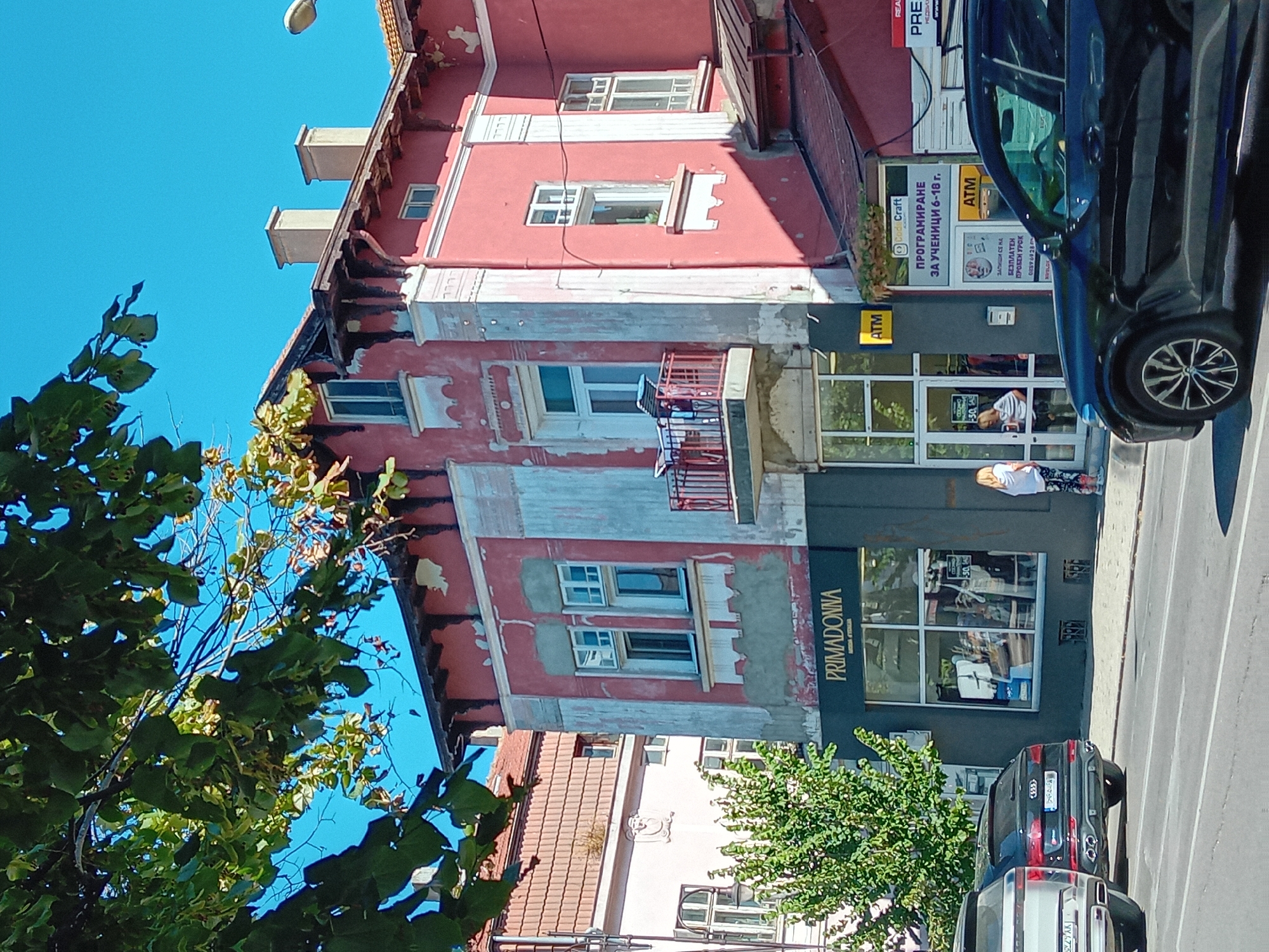 The image captures a street scene in the city of Varna, Bulgaria, on a clear, sunny day. The foreground features two luxury SUVs parked on the right side of the street, with a black Audi Q8 on the left and a silver Range Rover on the right. The street itself is asphalt with visible lane markings.
Dominating the mid-ground is a two-story building with a striking pink facade, showing signs of wear and peeling paint, particularly on the upper sections. The building features ornate architectural details, including decorative moldings around windows and a prominent balcony with a red railing. A dark-colored storefront, labeled "PRIMADONNA," is visible on the ground floor, with a large display window showcasing clothing. Next to it, a glass-fronted entrance leads into another establishment, identified by yellow ATM signage and a sign advertising "PROGRAMIRANE ZA UCHENICI 6-18 g." (programming for students 6-18 years old). A person, a woman with blonde hair, is standing by the entrance, appearing to interact with someone inside or look at the display.
In the background, another building with a tiled roof and lighter pink facade is partially visible. Lush green trees, with sunlight filtering through their leaves, frame the top left of the image, casting shadows on the scene. The clear blue sky forms the backdrop. The overall impression is that of a busy urban environment with a mix of residential and commercial properties.