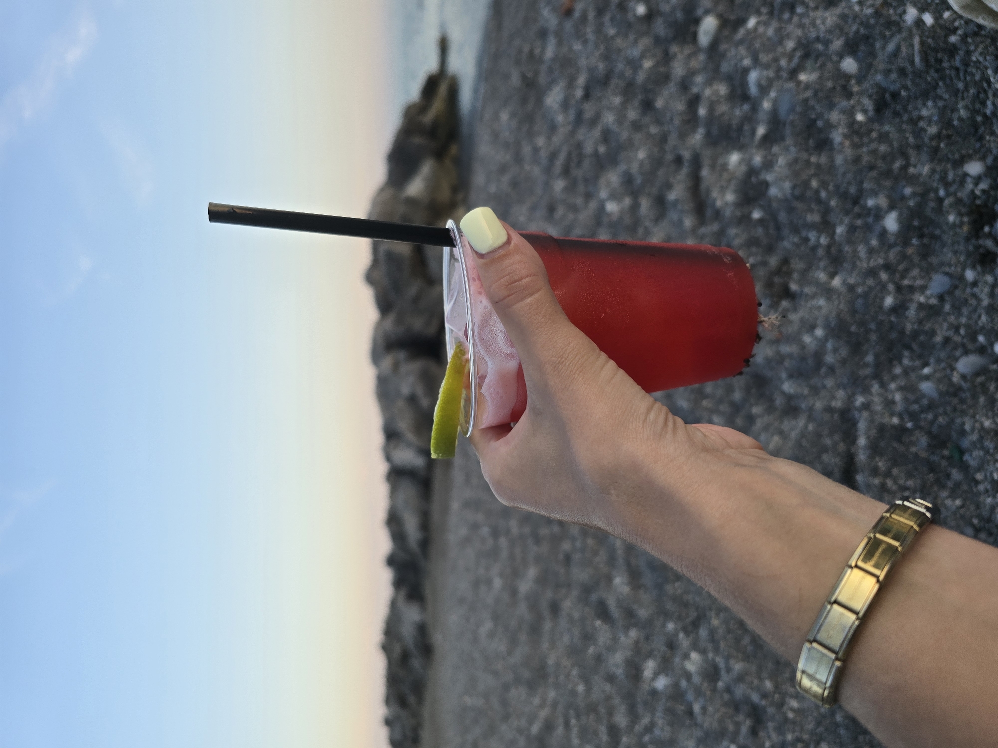 A person's hand, adorned with a gold bracelet and pale yellow nail polish, holds a clear plastic cup filled with a red beverage. A black straw extends from the cup, topped with a lime wedge. The cup rests against a pebbly beach, with a rocky coastline and the ocean visible in the background under a pale blue sky at dusk. The scene is not specifically identifiable as Salerno, Italy.