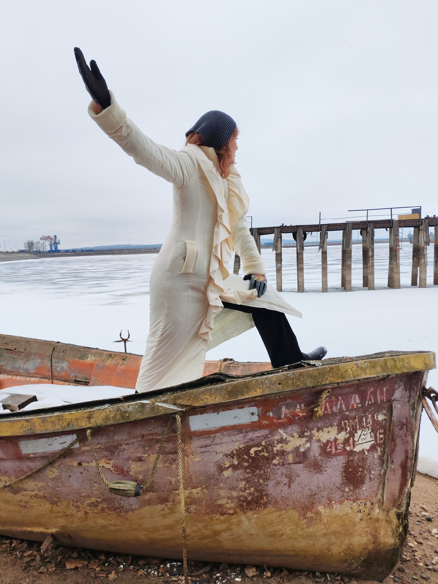 A woman stands in an old, weathered boat on a shore with a frozen body of water in the background. She is wearing a long, cream-colored coat with ruffles, black pants, a gray beanie, and black gloves. Her right arm is raised, as if gesturing towards the scene.
The boat is reddish-brown with peeling paint and some yellow trim along the top edge. There are ropes and a small wooden block attached to the side. Part of the side of the boat is marked with faded writing that includes Cyrillic letters and some numbers. Snow is visible inside the boat.
The background consists of a frozen body of water, possibly a river or lake, covered in ice and snow. A wooden pier or structure with evenly spaced pilings extends into the water. Further in the distance, there is a hazy sky, and some industrial structures, including cranes and other buildings, can be seen. This is Chaikovsky, Russia, based on identifying features in the scenery.