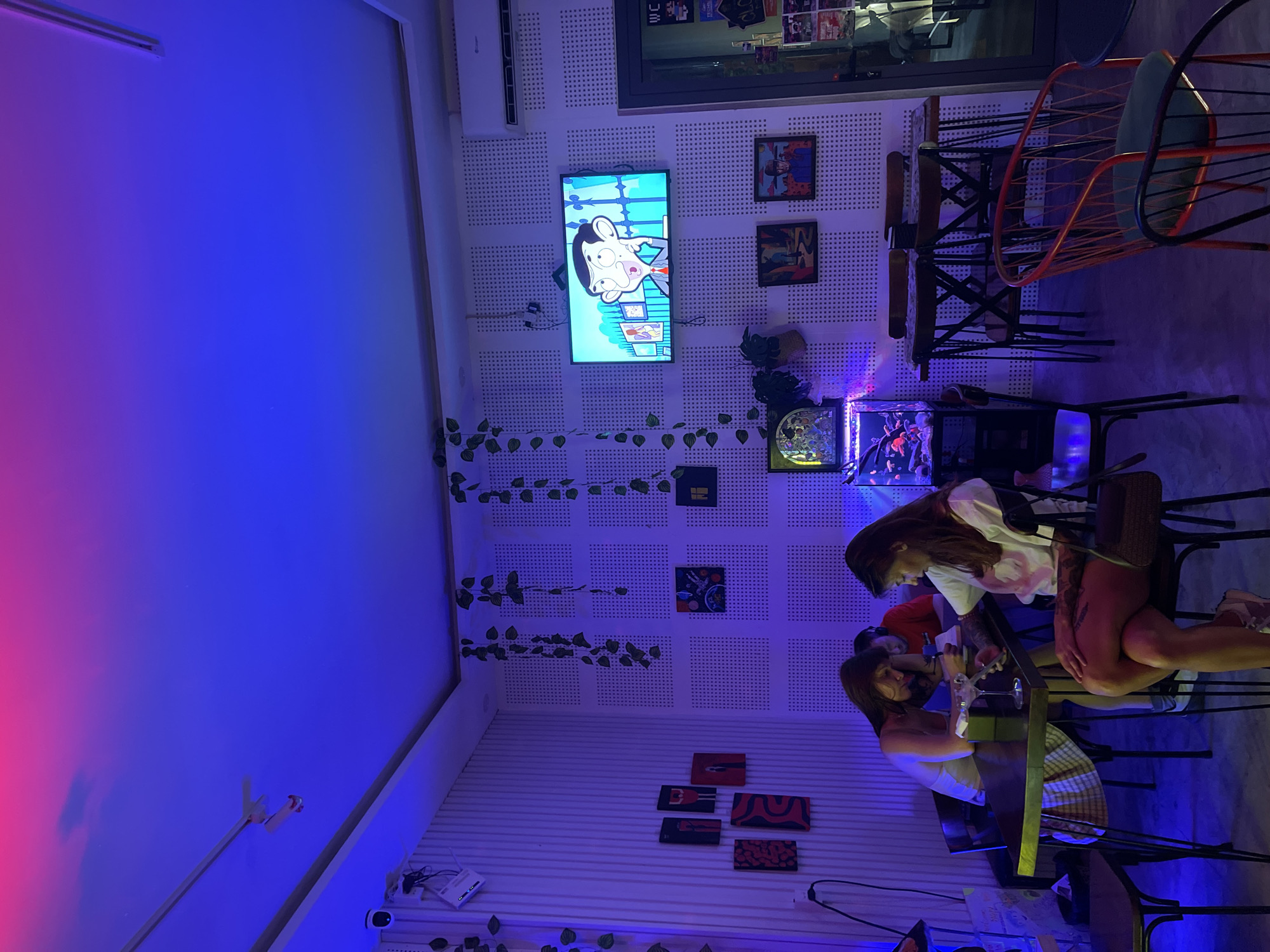 The image depicts the interior of a dimly lit establishment, likely a cafe or bar, with a pronounced blue and pink neon lighting scheme illuminating the space. The walls are white and textured, featuring a grid of small perforations. Several framed pictures and artwork are displayed on the walls. Green artificial vines hang from the ceiling, adding a touch of greenery to the decor.
In the foreground, to the right, a red metal chair with a dark seat is visible. The environment is furnished with various tables and chairs, some of which are occupied. In the mid-ground, two women are seated at a table. One woman, on the left, is wearing a white top and a checkered skirt, leaning over the table and appearing to be engaged with her phone. The other woman, on the right, is wearing a white t-shirt and is also looking at her phone, with her arm resting on the table. Another person, a man wearing a red t-shirt, is seated behind them, looking down at something on the table.
On the wall above the seated individuals, a television screen displays an animated character, resembling Mr. Bean. To the left of the TV, a white air conditioning unit is mounted. In the lower left corner, a table with a glass and what appears to be a drink is partially visible. Further to the left, a security camera is mounted on the wall. In the far left of the image, a black chair is present. In the mid-ground, to the right of the seated patrons, there are two small tables with chairs around them, and a fish tank with visible fish.
The scene is characterized by its cool, ambient lighting and a relaxed, social atmosphere. The presence of artwork, a TV, and comfortable seating suggests it's a place for leisure and entertainment. The location is identified as being in the city of Da Nang, Vietnam.