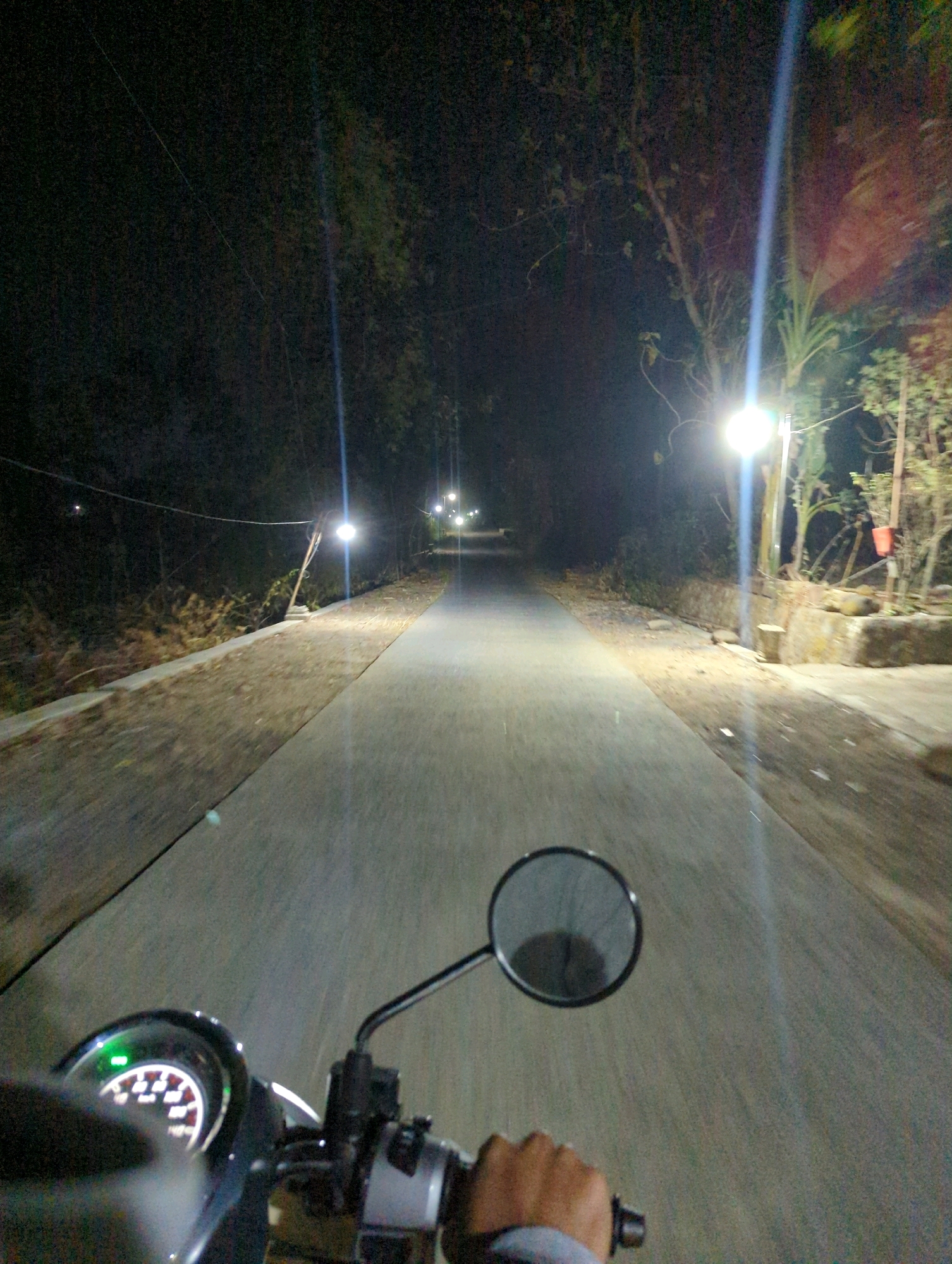 The image depicts a first-person perspective from a motorcycle traveling down a paved road at night. Streetlights illuminate the scene, casting light on the road and the surrounding vegetation. The road appears to be in a rural or semi-rural area, with trees and foliage visible on either side. In the foreground, the motorcycle's handlebars, a rearview mirror, and a portion of the dashboard are visible. The dashboard shows a speedometer with a green indicator light. The location is specified as Kedungdowo, Indonesia.