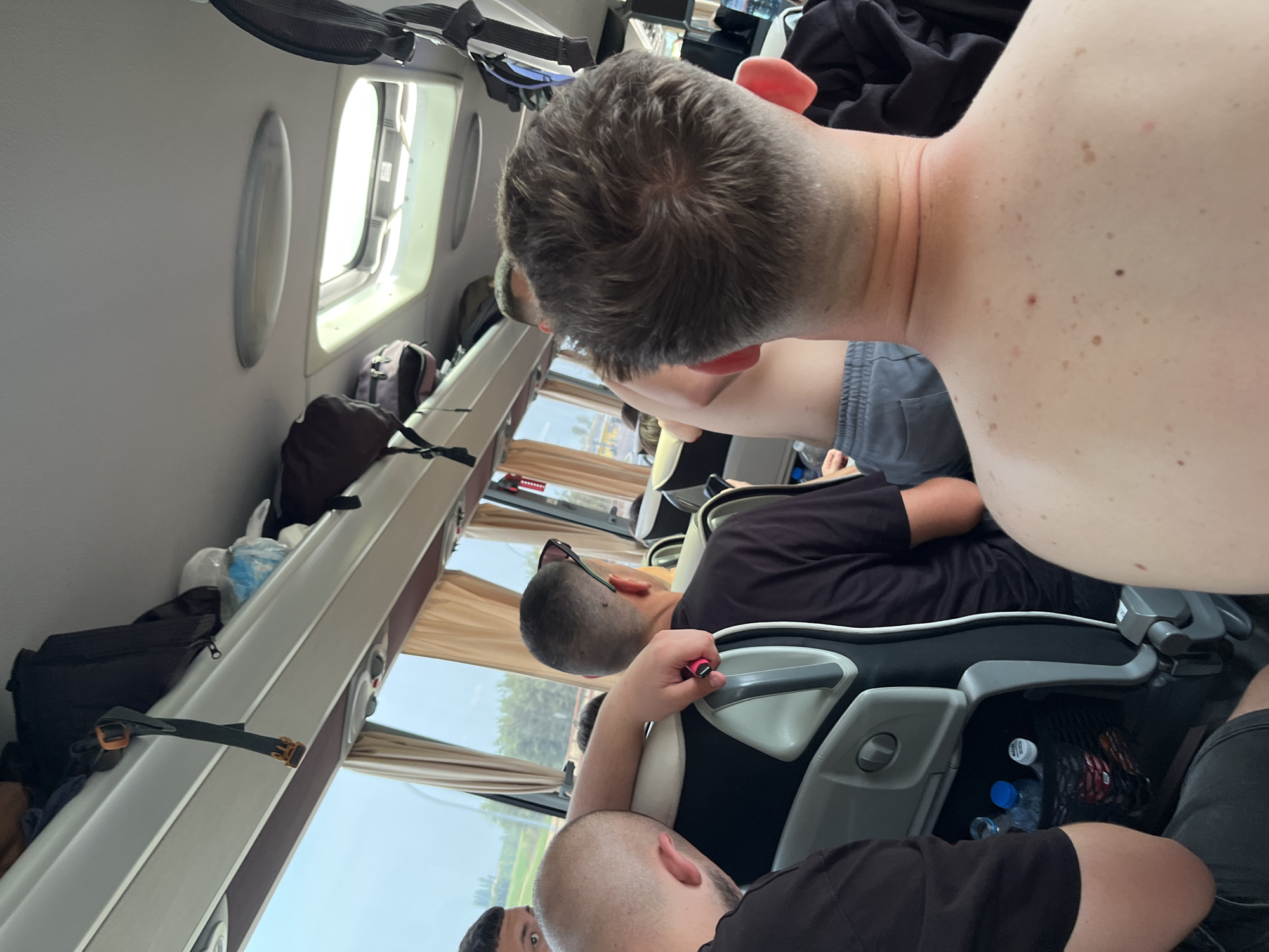 The image shows the interior of a passenger vehicle, likely a bus or private jet, with multiple people seated. Natural light streams in through the windows. Overhead storage compartments are visible. Passengers are wearing casual clothing. One man is shirtless. Water bottles are stowed next to a seat. The location is implied to be near Bottega Panzano, Italy, based on the request context but there is nothing in the image to confirm this.