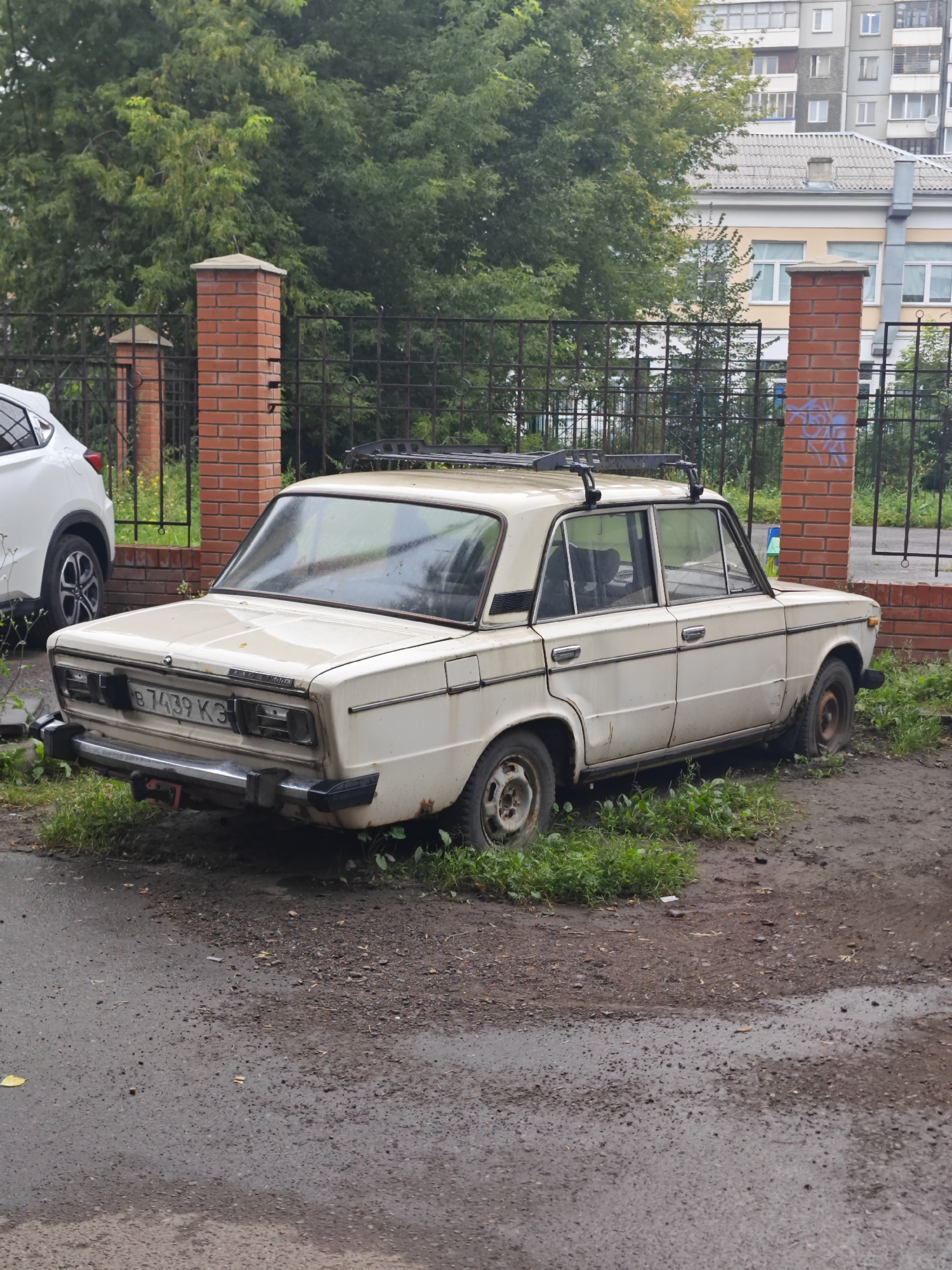 A beige, older model sedan with a roof rack is parked on an unpaved area beside a paved road, surrounded by overgrown grass and weeds. The car has a flat rear tire on the passenger side. A chrome bumper is visible on the rear of the car, and it has a license plate that reads "B 7439 K3". A portion of a white SUV is visible on the left side of the image, parked behind a brick fence. Behind the fence, trees and buildings are visible. The scene appears to be in a residential area.