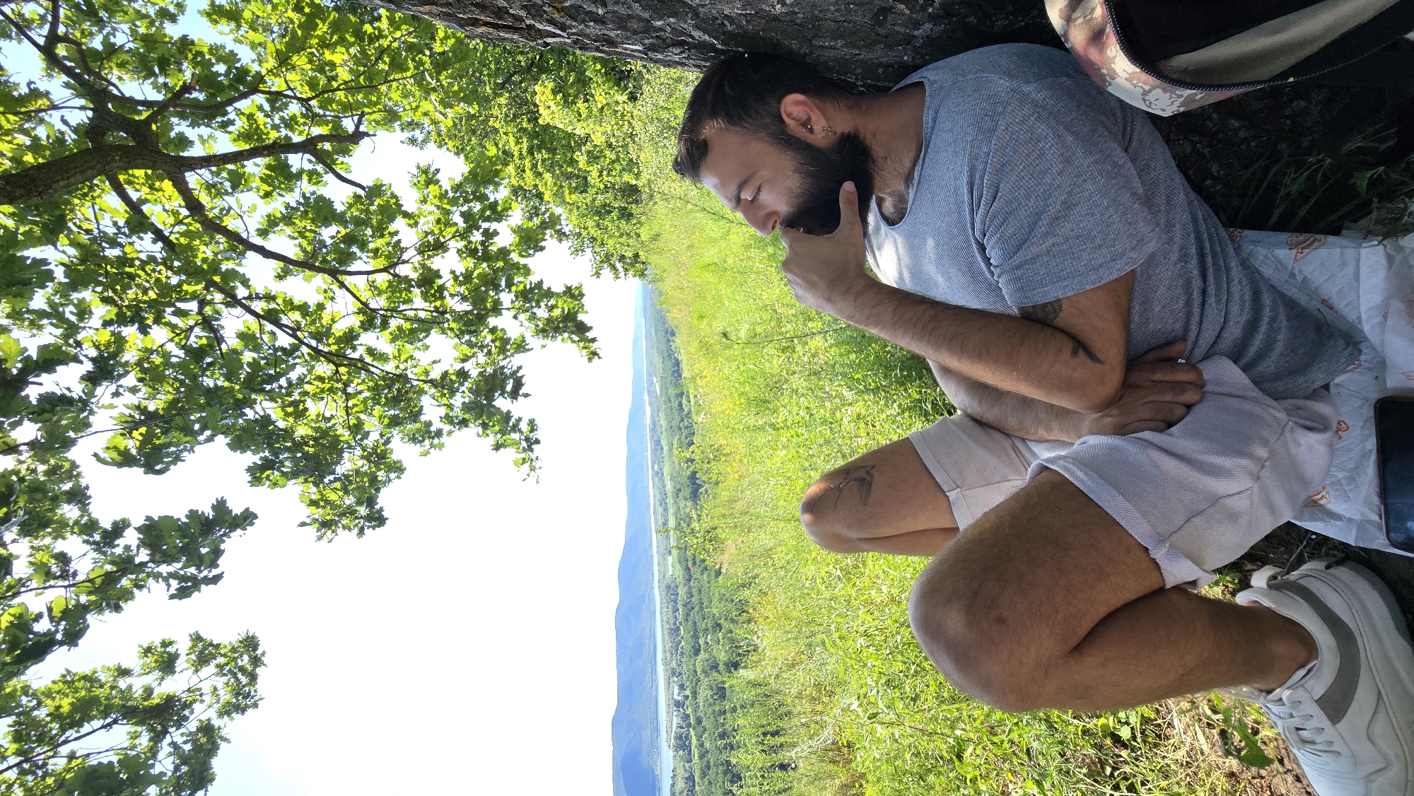 A man with a beard and short hair is resting against a tree on a grassy hillside overlooking a valley. He is wearing a grey t-shirt and white shorts. His right hand is resting on his chin, and his eyes are closed. In the background, a wide river flows through a green, wooded valley, with hills in the distance. The sky is bright and clear. The scene appears to be set in a natural environment, possibly a park or a nature reserve, consistent with a location in the city of Samara, Russia, known for its picturesque Volga River landscapes.