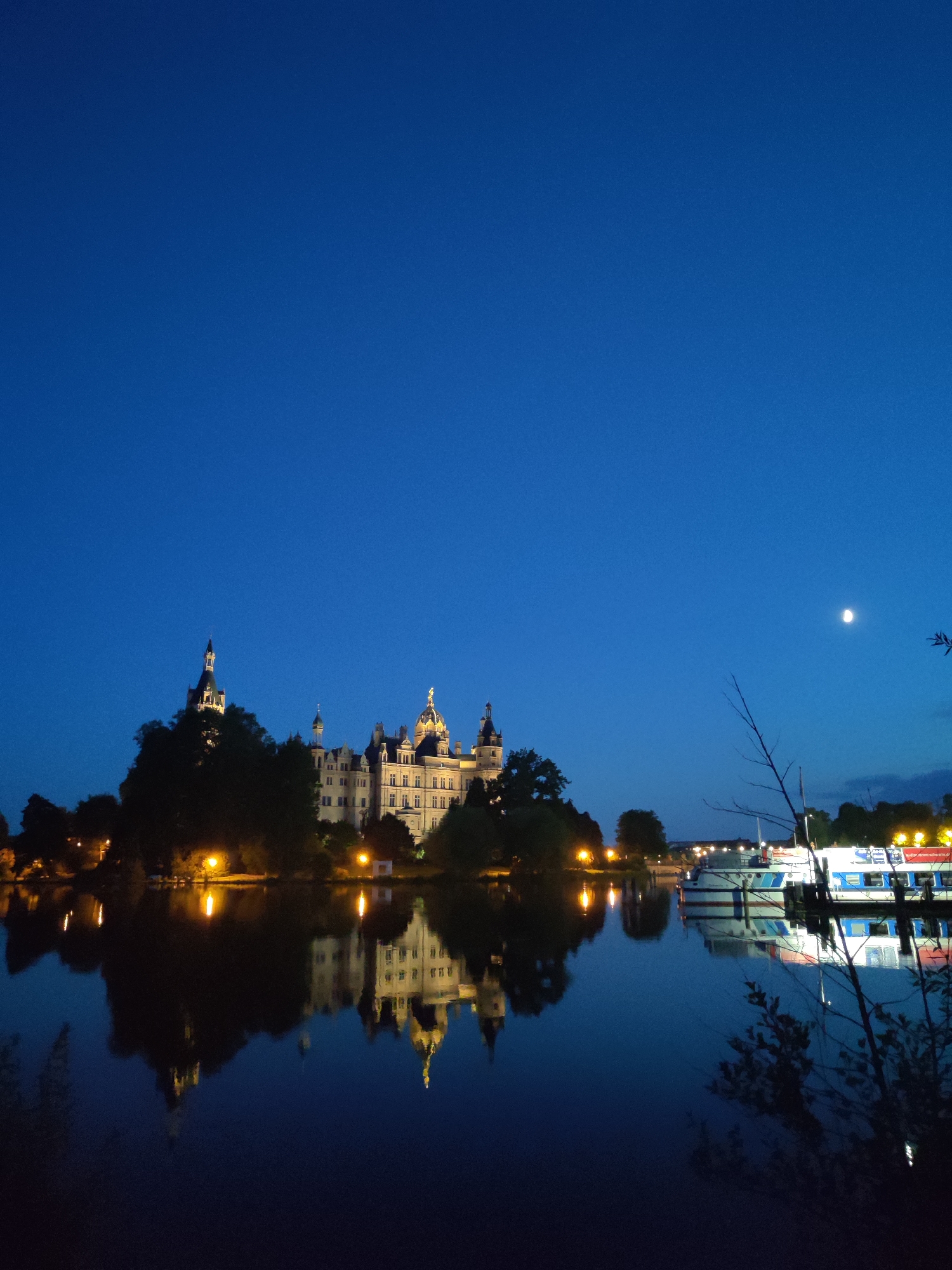 This is a nighttime vertical photograph of Schwerin Castle, located on an island in the Schwerin Lake in Schwerin, Germany. The castle is illuminated by artificial lights and is reflected in the still water. The sky is a deep blue with a sliver of the moon visible in the upper right quadrant. Several boats are docked to the right of the castle. Trees are visible in front of the castle, some of which are also illuminated by streetlights.