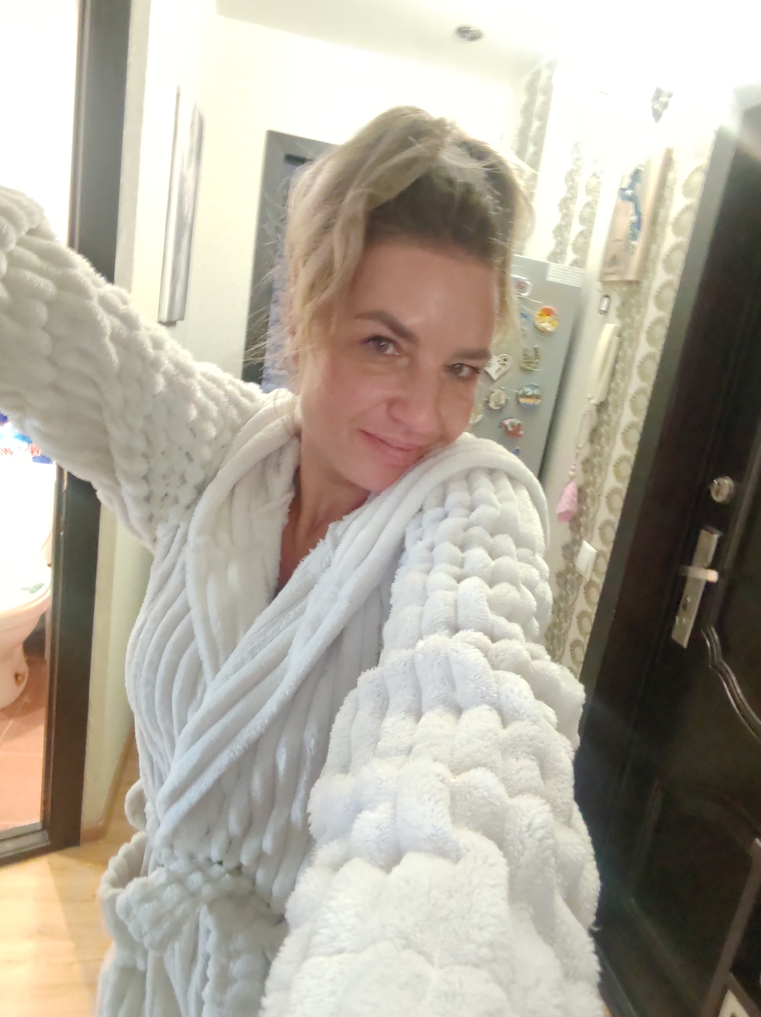 A woman takes a selfie indoors, likely within a residence in Petropavlovsk-Kamchatsky, Russia. She smiles softly at the camera, wearing a plush, white bathrobe with a distinct textured pattern on the sleeves and body. Her light-colored hair is pulled back, visible on the top of her head.
The background offers a glimpse into a typical home environment. To the left, part of a bathroom with a toilet and dark door frame is visible. To the woman's right, a refrigerator covered with numerous magnets stands against a wall adorned with patterned wallpaper. Above the refrigerator, a light fixture is seen on the ceiling. Further to the right, a dark brown door, possibly an entrance door, with a silver handle and lock is prominent. A wall-mounted intercom or phone is situated above the door handle, and a decorative item, possibly a wooden plaque, hangs on the wall. The flooring appears to be light-colored wood or laminate. The lighting suggests artificial indoor illumination. The scene conveys a casual and relaxed atmosphere. No legible text is visible in the image.