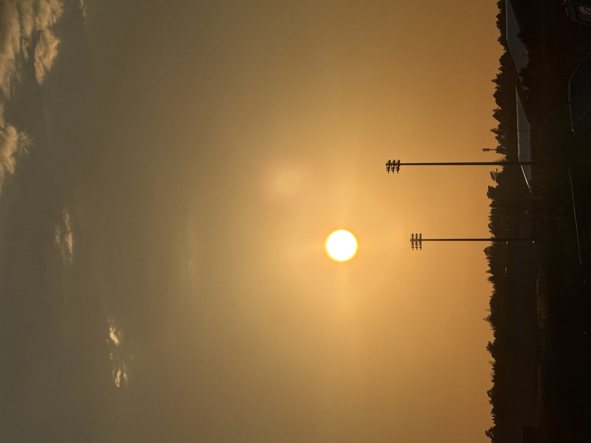 The image captures a sunset over a rural area, likely on the outskirts of Galinė, Lithuania. The sky is a dominant orange hue, with a bright sun positioned slightly left of center. Faint, wispy clouds are visible in the upper portion of the sky. In the mid-ground, silhouetted against the sunset, are two tall, slender poles topped with multi-tiered lighting fixtures, resembling stadium or sports field lights. These poles stand out against a dark, tree-lined horizon. Below the horizon, the landscape is largely obscured by shadow, but hints of structures, possibly buildings with sloped roofs, can be discerned. The foreground is also dark and appears to be a ground-level view with indistinct shapes that could be vehicles or other objects. There are no people visible in the image. The overall impression is one of dusk settling over an open, potentially recreational, area.