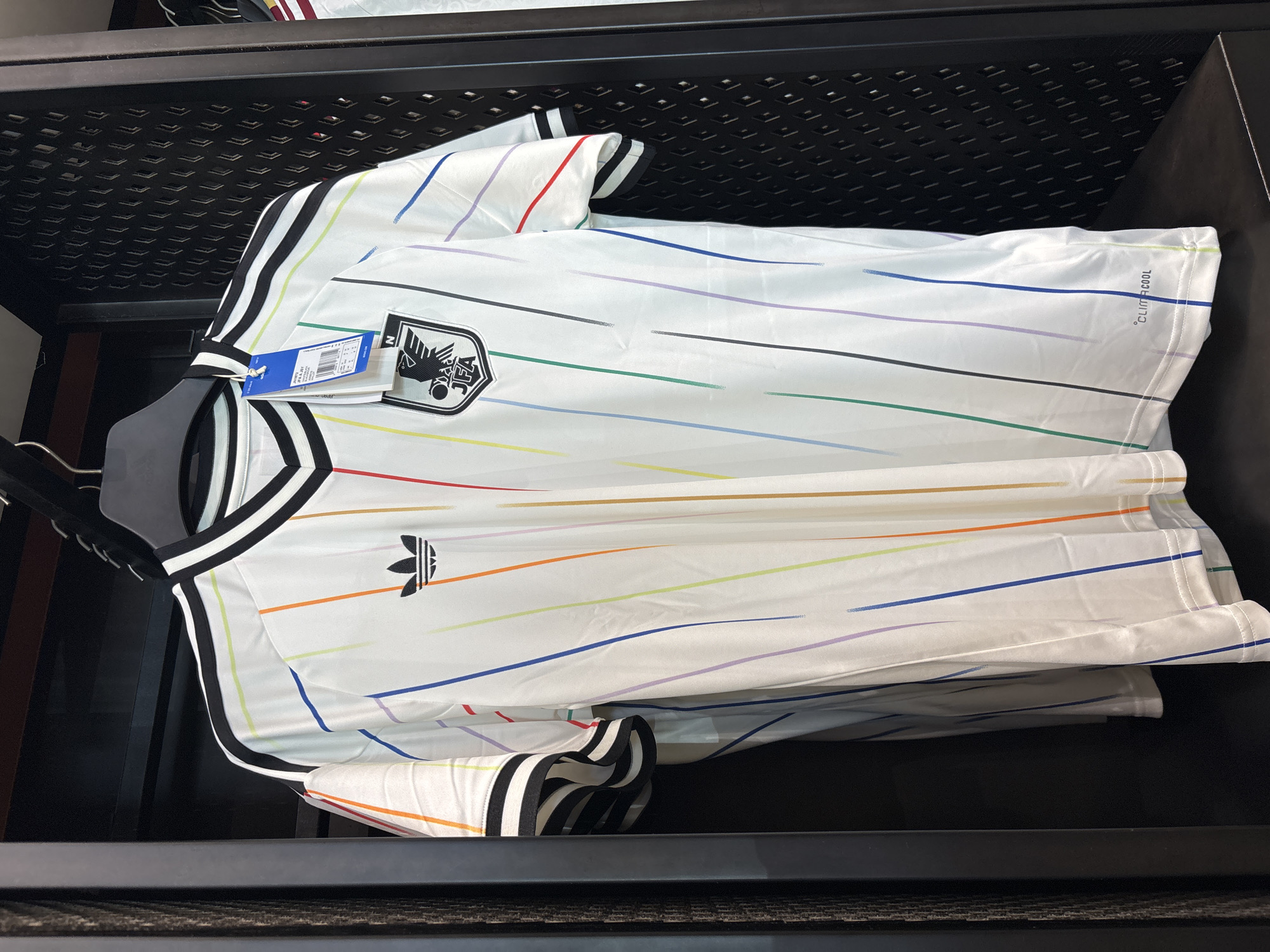 A white Adidas soccer jersey with colorful stripes is folded and placed on a hanger within a black, grated shelf. The jersey has the Adidas logo and the Japan Football Association (JFA) crest. A blue Adidas tag is visible on the hanger. The shirt is labeled as “climacool”. The image appears to have been taken indoors, possibly in a store. There is no evidence of the city of Sao Paulo, Brazil.