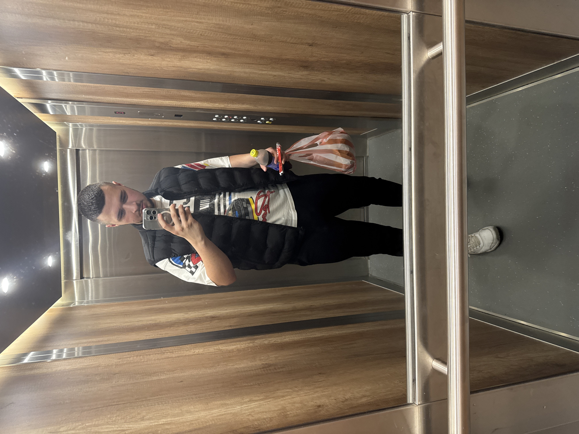 A young adult male is standing in an elevator and taking a selfie. He is wearing a black puffer vest over a white t-shirt with a graphic print, black pants, and white sneakers. He is holding a smartphone in his right hand and a plastic bag with food and a bottle of soda in his left hand. The elevator has wood-paneled walls and stainless steel accents. The floor is dark gray with a speckled pattern. The lighting is bright, coming from overhead fixtures. The elevator appears to be located in an apartment building in Sofia, Bulgaria.