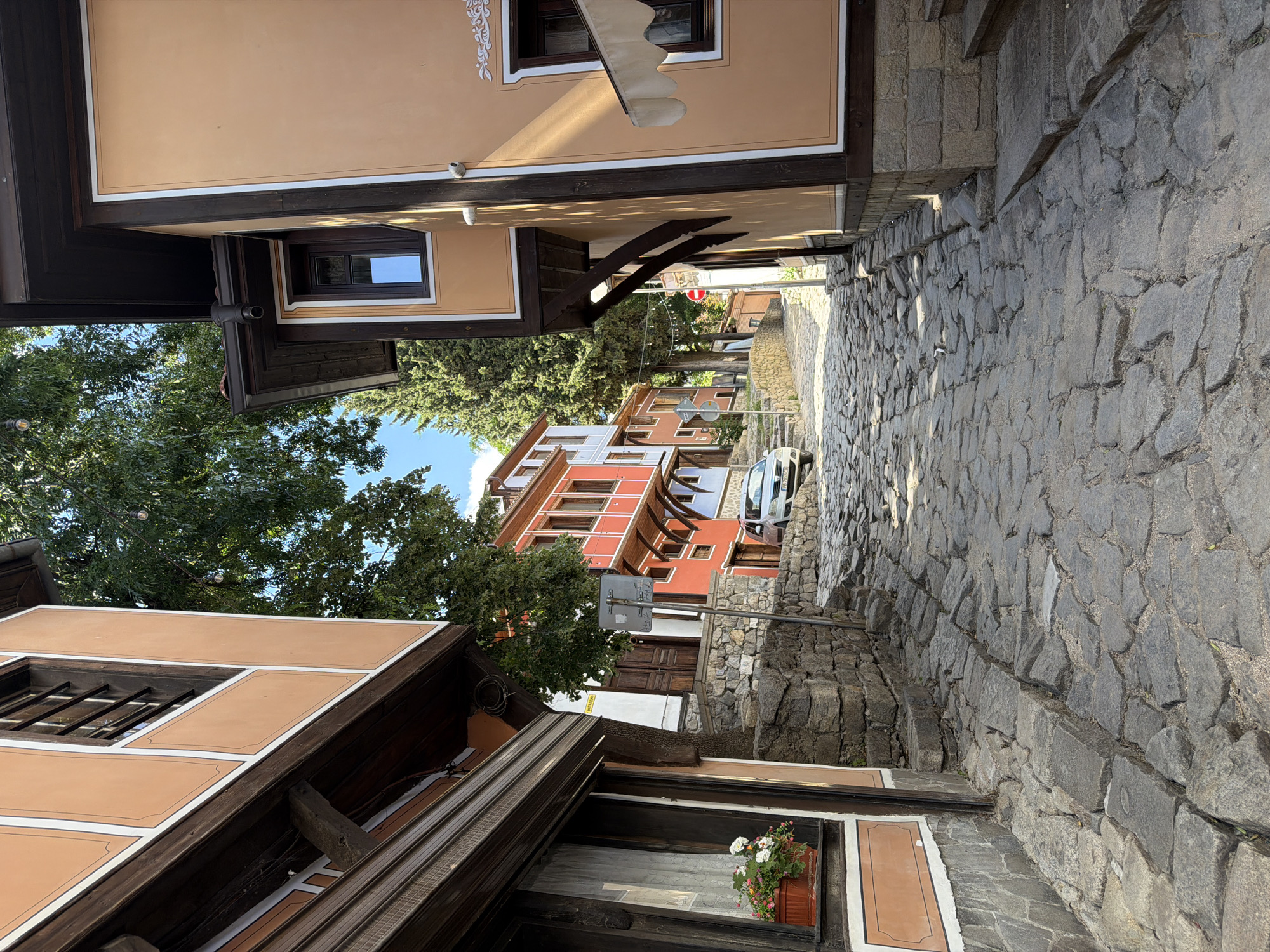 The image shows a narrow, cobbled street in Plovdiv, Bulgaria, lined with traditional Bulgarian Revival-style houses. The houses are painted in warm colors, with wooden architectural details. Trees are visible, creating a partial canopy over the street. A white car is parked further down the street. The street is angled upwards. The buildings on either side of the street are closely packed together. A stone wall is on the right side of the image. A window with a flower box is visible in the foreground. A street sign is visible in the mid-ground.