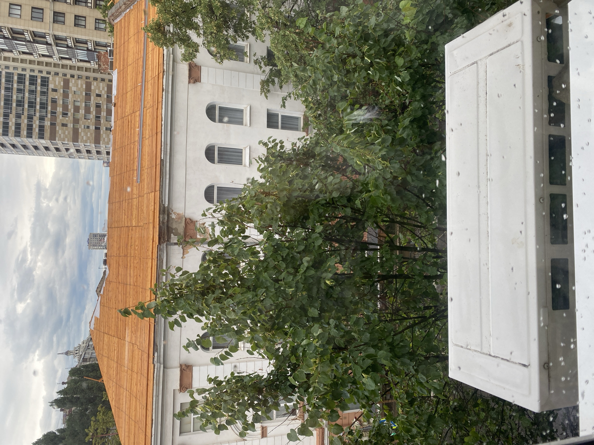 The image captures an exterior view of a building under renovation, likely in Novaya Usman', Russia, as suggested by the context. The foreground features the top of a white air conditioning unit, partially obscured by raindrops on a window pane. Beyond the unit, lush green trees with dense foliage dominate the mid-ground. Behind the trees, the upper floors of a multi-story building are visible, featuring a light-colored facade with arched windows and decorative architectural elements. The roof of this building is covered with an orange material, possibly wooden planks, indicating ongoing construction or repair work. In the background, taller, modern buildings rise against a cloudy sky, with a spire-like structure visible to the left, further suggesting an urban or suburban setting. The overall atmosphere is overcast, with visible raindrops on the glass in the foreground contributing to a damp or rainy day impression. There are no people visible in the frame.