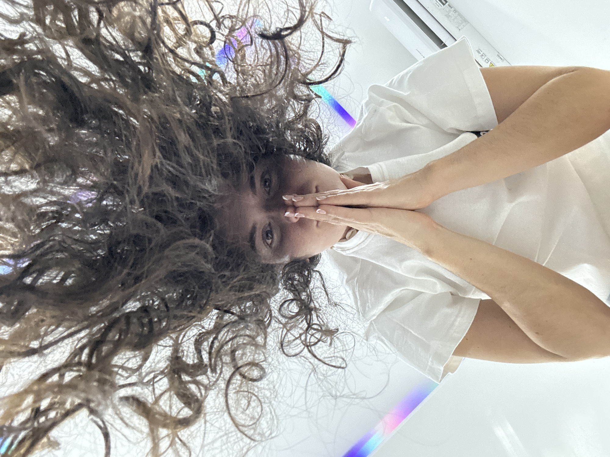 This overhead portrait features a person with voluminous, dark, curly hair, spread out against a bright white background. The subject is wearing a white short-sleeved t-shirt. Their hands are clasped together, covering their mouth and nose, with light-colored manicured fingernails visible. Only their dark, expressive eyes are fully visible, looking directly at the viewer.
The setting appears to be an indoor space, dominated by clean, bright white surfaces, likely walls or a floor. The lighting is very bright, creating subtle shadows within the curly hair. A soft, iridescent rainbow-like light streak is visible across the white background and within the hair, suggesting a reflection from a prism or similar effect. In the upper right corner, part of a white wall-mounted air conditioning unit or similar appliance is visible.
The activity is a posed portrait, with the hands covering the face conveying a sense of shyness, contemplation, or a quiet gesture. The unusual high-angle perspective, with the subject seemingly lying down or positioned below the camera, creates an intimate and artistic composition. While the location context is Mũi Né, Vietnam, no specific geographical cues are discernible within the image itself beyond the presence of an air conditioner. No readable text is clearly visible.