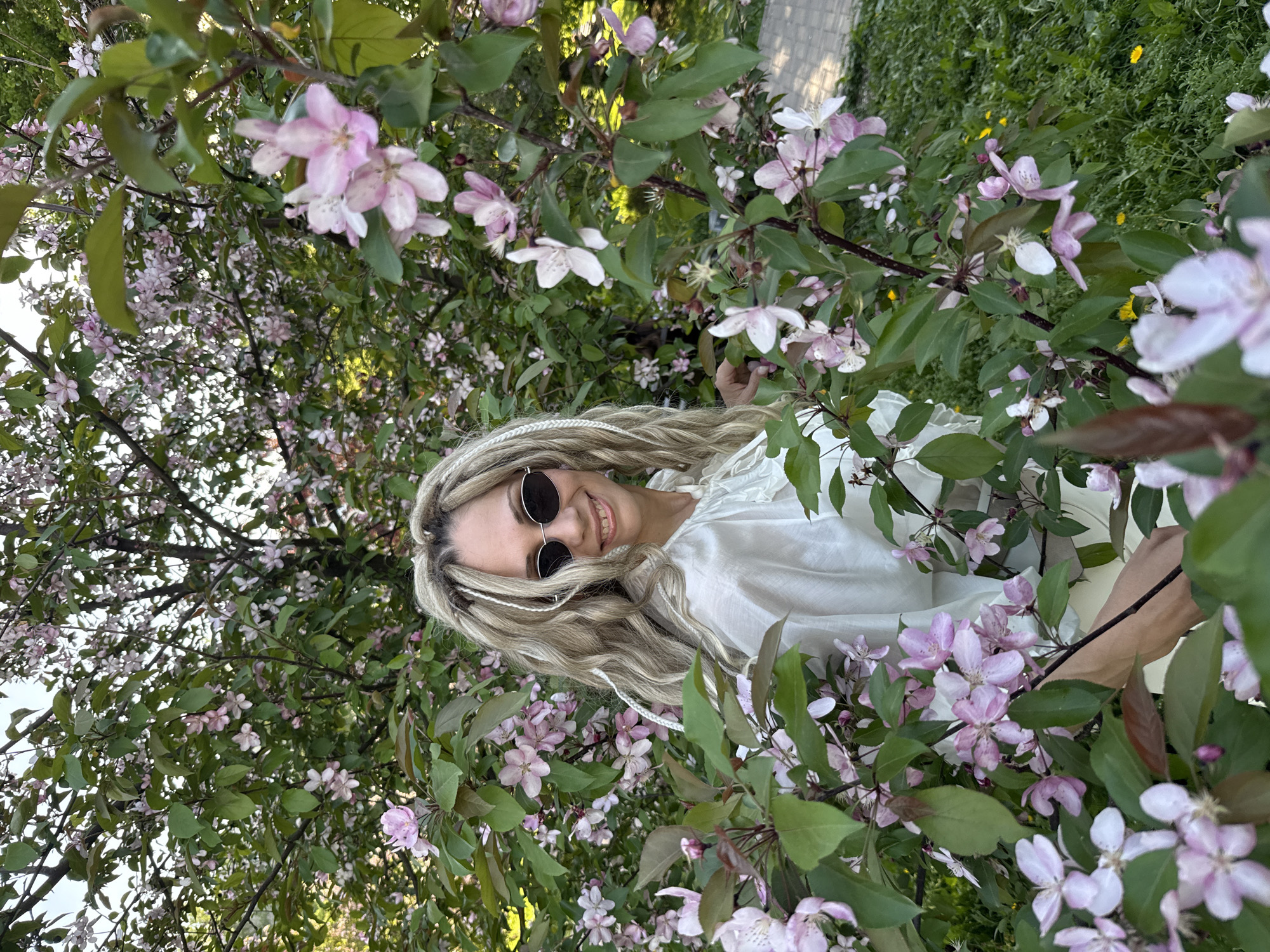 A woman with blonde hair styled in dreadlocks and wearing sunglasses and a white blouse stands among the branches of a flowering tree with pink and white blossoms. She is smiling and looking directly at the camera. The tree is densely covered in leaves and flowers, and some grass and a paved path are visible in the background. The location appears to be in a park or garden in Rusanovo, Russia.