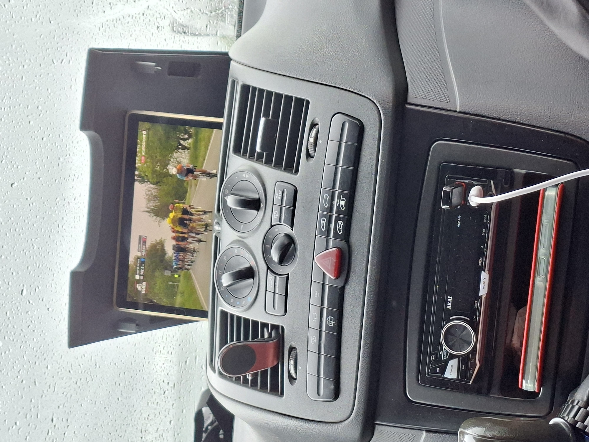 The image shows the interior of a vehicle, specifically the dashboard area. A tablet is mounted on the dashboard displaying a video of a cycling race. The vehicle's air vents, climate control knobs, hazard light button, and other control buttons are visible. To the right of the climate controls, there is an AKAI car stereo and a smartphone, with a charging cable plugged into the stereo. The location, Dragijevo, Bulgaria, is not evident in the visual details.