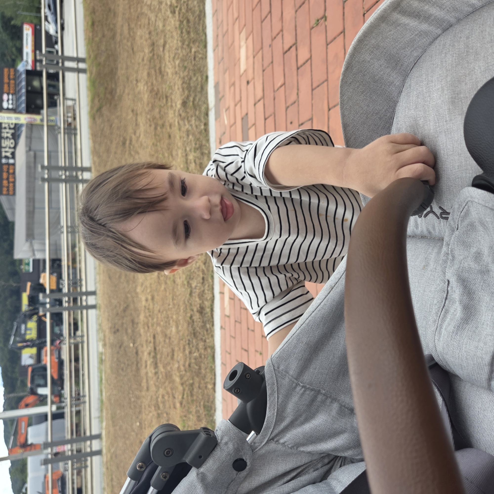 In Dunnae-myeon, South Korea, a young child with light brown hair is shown in the foreground, wearing a black and white striped t-shirt. The child is leaning forward, looking down and to the left with their tongue slightly out. The child's right hand is reaching towards a grey stroller. The stroller is a prominent object in the image, with its fabric covering and black handle visible. The background consists of dry, brown grass, a brick-paved pathway, and a blurred scene of construction equipment, including excavators, and buildings. The overall setting appears to be an outdoor area, possibly a park or a rest stop, adjacent to a roadway.