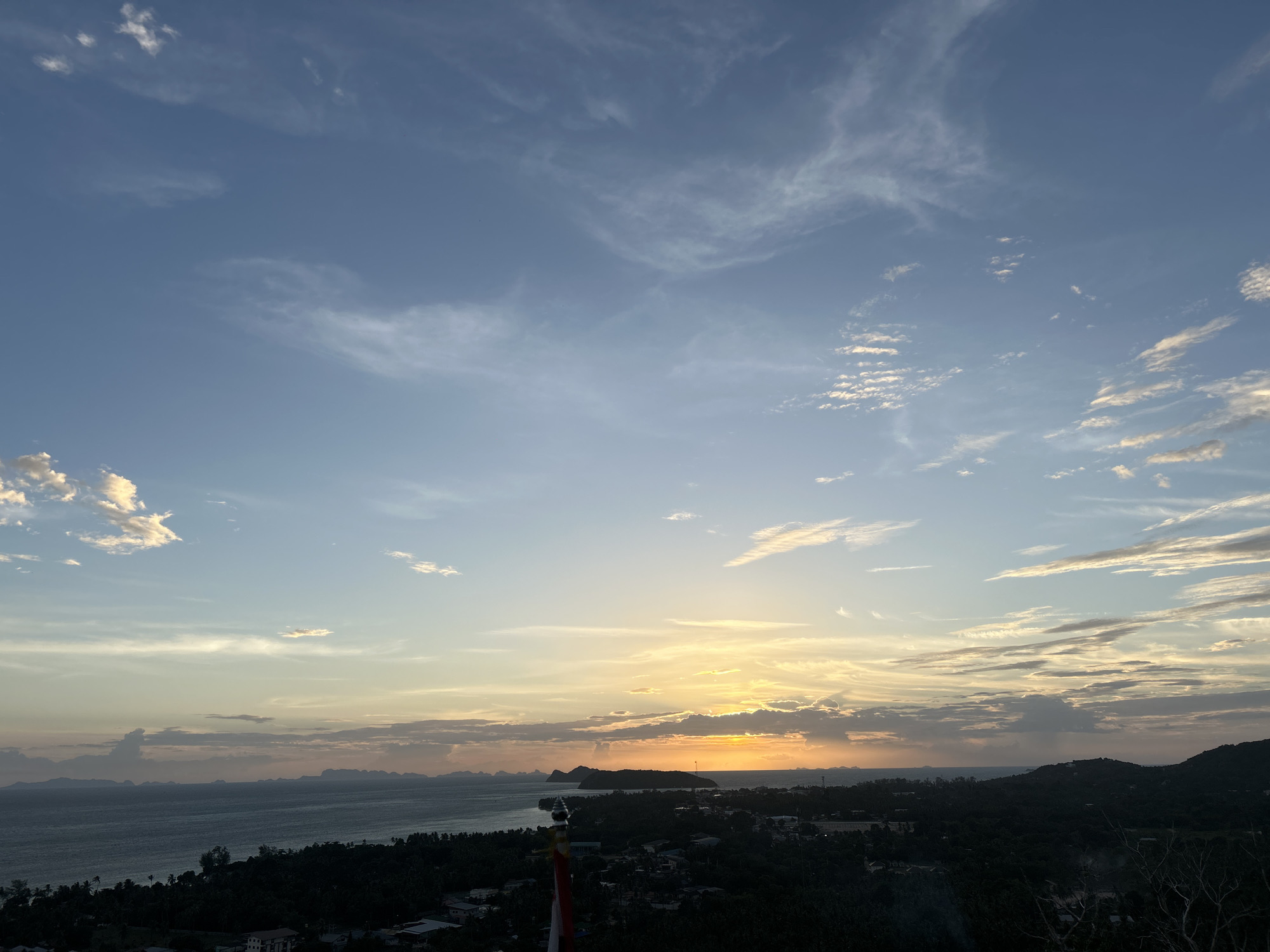 A scenic aerial shot captures the sunset over the Gulf of Thailand, viewed from Ko Pha-ngan District, Thailand. The sky is painted with a gradient of colors, from a clear blue at the top to soft oranges and yellows near the horizon. Wispy clouds streak across the sky, catching the last rays of the sun. Below the sky, the vast expanse of the ocean stretches out, reflecting the sky's colors. The coastline is dotted with lush green trees and small buildings, indicating a coastal settlement. Islands are visible in the distance, silhouetted against the colorful sky. The foreground is dominated by the dark silhouettes of trees and vegetation, hinting at the elevated viewpoint from which the photograph was taken. In the lower right corner, a faint structure or object is visible, partially obscured by darkness.