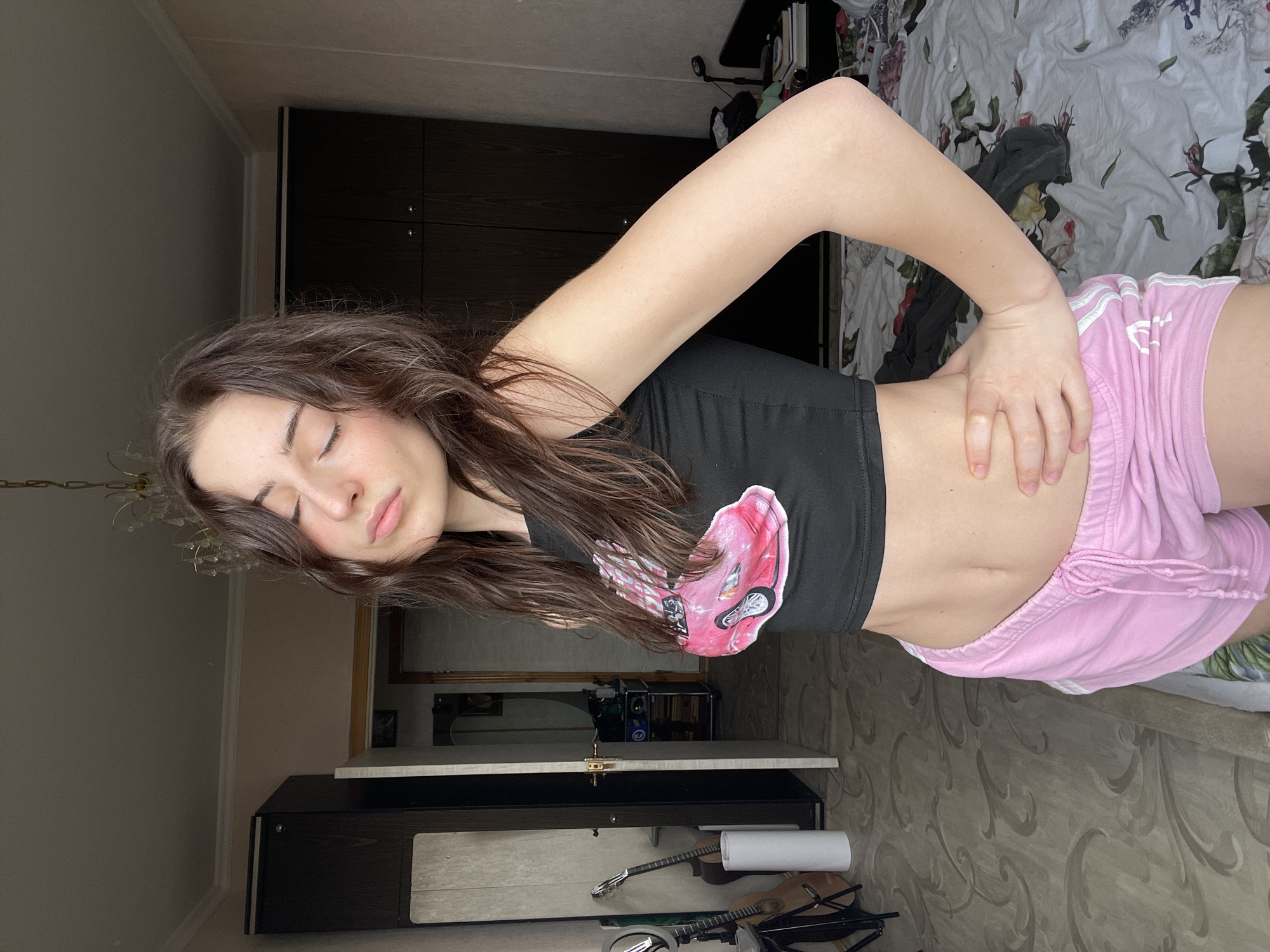 A young woman with long brown hair and fair skin stands indoors, wearing a black crop top with a pink design and pink shorts with white stripes. She has her eyes closed and her left hand is on her hip. The background includes a bed with a floral pattern, a dark wardrobe, and a decorated floor. The location cannot be confirmed as Moscow, Russia based on the visible elements. The scene appears to be in a bedroom.