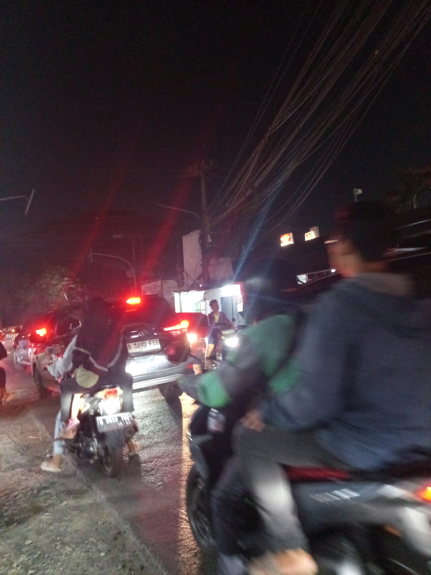 The image captures a nighttime scene in Karangsatria, Indonesia, depicting a traffic jam on a wet road. In the foreground, a person is seated on a motorcycle, facing away from the camera, with their legs visible. Another motorcycle is positioned to the right, partially obscured. In the mid-ground, a car with illuminated taillights and a license plate reading "B 1695 FIS" is visible. Additional vehicles and people are present in the background, indicating a busy urban environment. Overhead, a dense network of power lines stretches across the dark sky, with a faint glow from buildings and streetlights illuminating the scene. The overall impression is one of a typical congested road at night in an Indonesian city.
