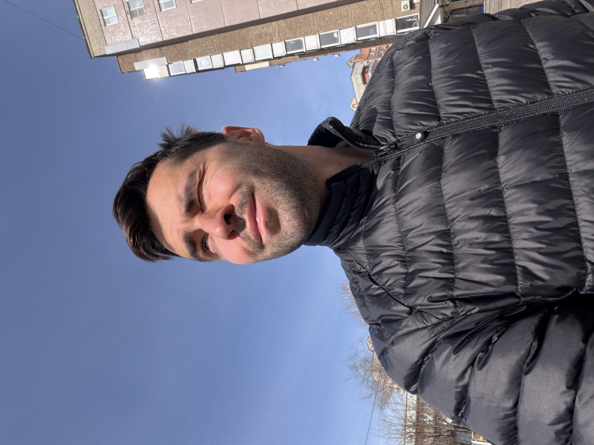A close-up, rotated portrait captures a man wearing a black puffer jacket outdoors. The sky above him is a clear, light blue. Behind him and slightly above is a multistory building with numerous windows, suggesting an urban environment. Considering the instructions specify the location as Krasnoyarsk, Russia, the picture was likely taken there. The man is looking directly at the camera with a slightly quizzical expression.