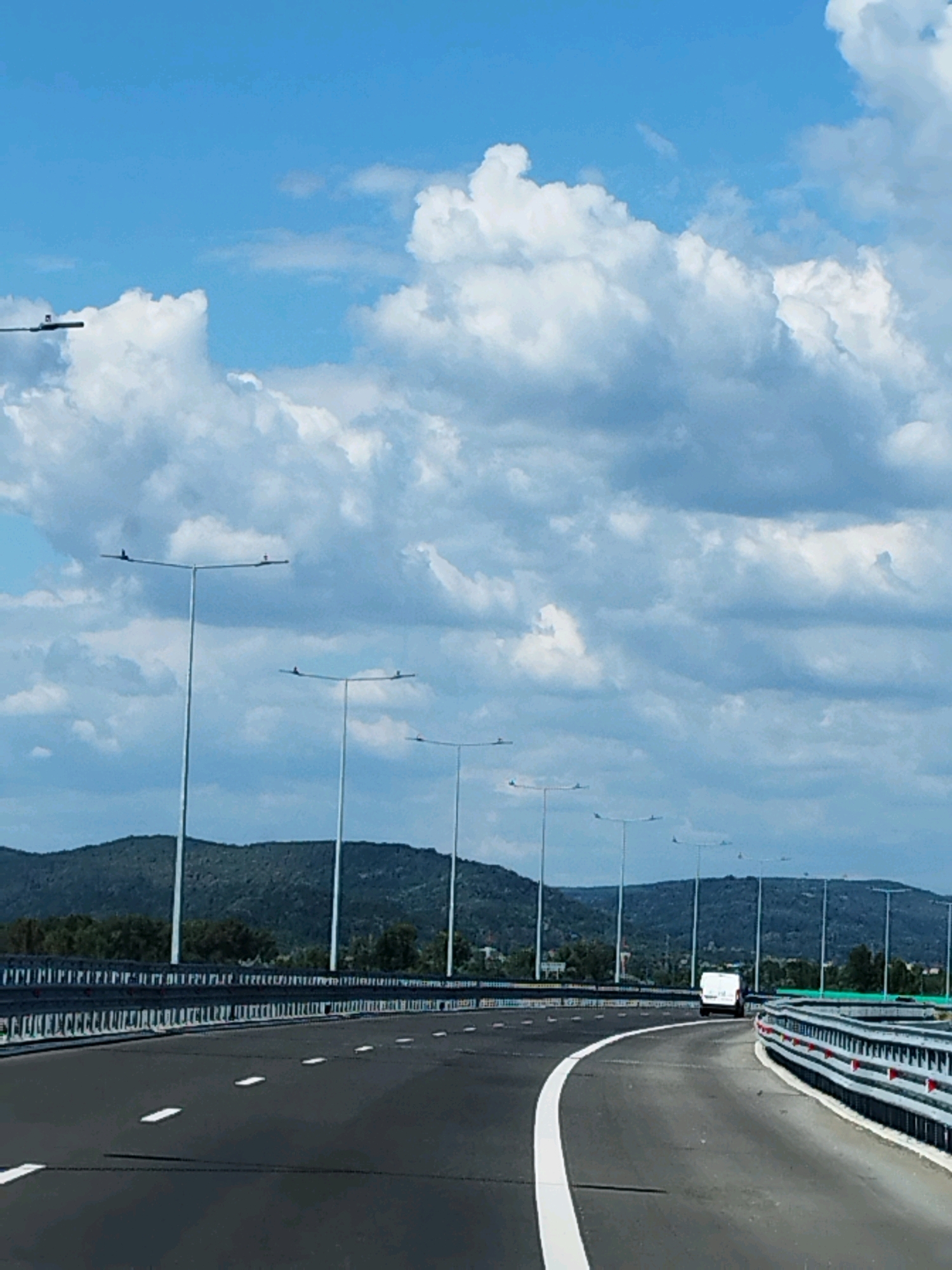 The image depicts a highway scene in Merișani, Romania. The perspective is from a vehicle traveling on the road, looking forward and slightly to the left. The road surface is asphalt with white lane markings. On the left side of the road, there is a metal guardrail and then a row of tall lampposts. In the distance, there are green, tree-covered hills under a blue sky with scattered white clouds. A white van is visible driving ahead on the road. The scene is daytime.