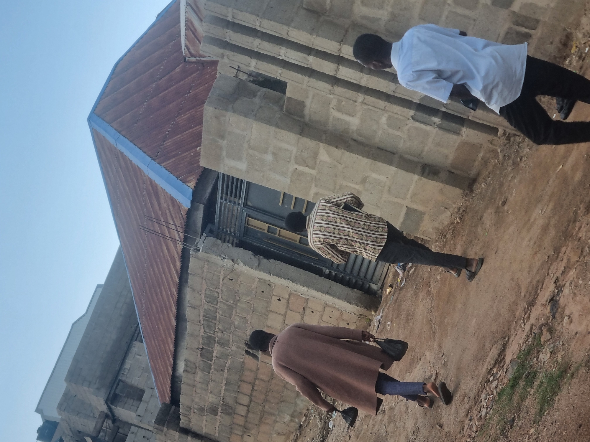 This media file shows a scene outdoors in Jos, Nigeria, likely during the daytime with clear skies. Three individuals are visible, all appearing to be male and walking away from the camera.
The first individual, closest to the camera, is wearing a brown overcoat and blue pants, carrying a black bag in their right hand. The second person, further back and to the left, is dressed in a patterned shirt with horizontal stripes in cream, brown, and black, along with dark pants and dark sandals. The third person, most distant and to the right, is wearing a white short-sleeved shirt and dark pants.
The setting appears to be an area with unfinished or partially constructed buildings. The building on the left has a corrugated metal roof, possibly rusted, with blue trim. The wall of this building is constructed from concrete blocks. Another building, to the right, is also made of concrete blocks. The ground is predominantly dirt and sand, with some scattered debris and patches of sparse vegetation. The overall impression is of an urban or semi-urban environment in a developing area. No visible text is present in the image. The scene is neutral in terms of expressed emotion, focusing on the movement of the individuals through the environment.