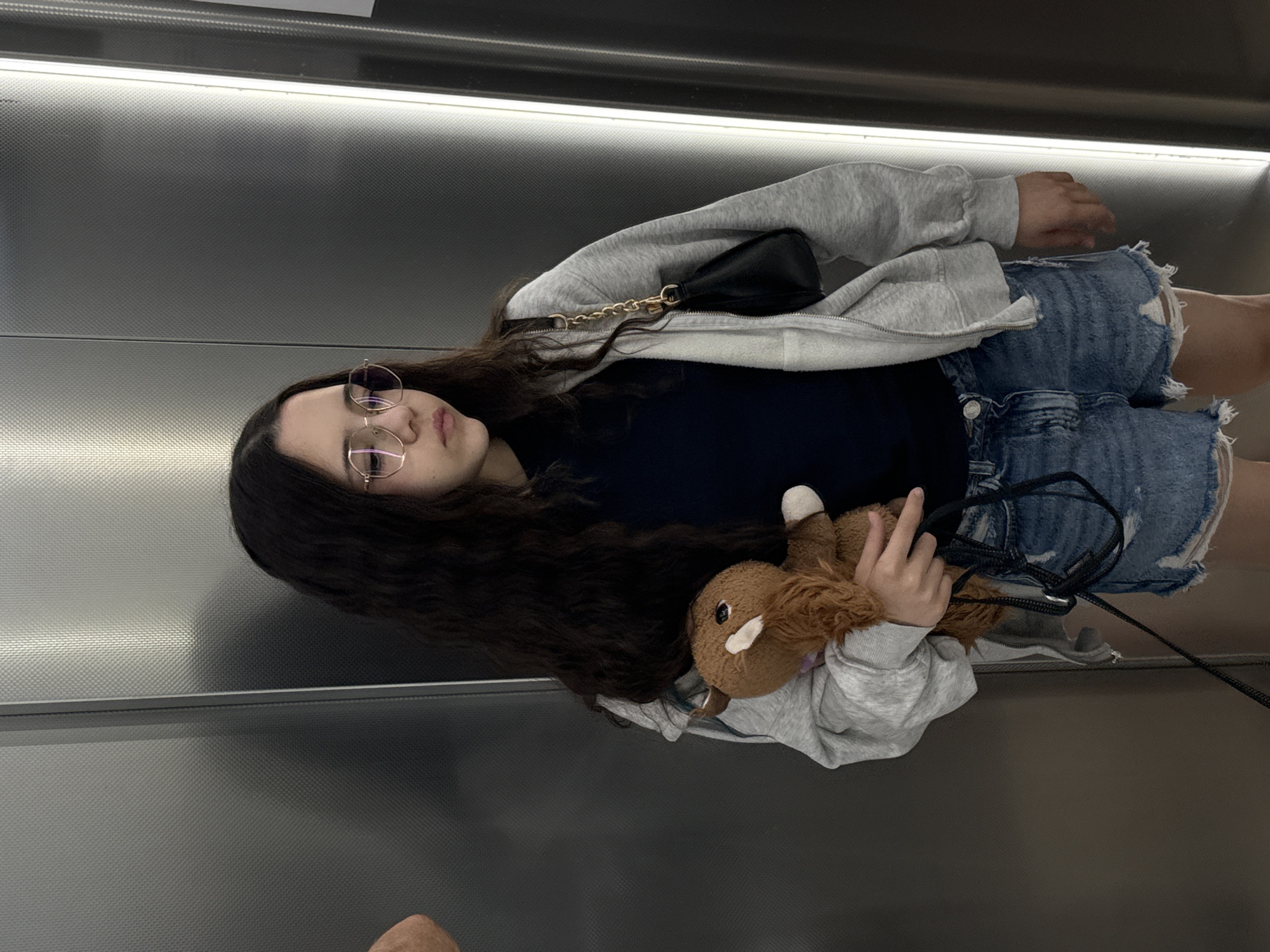 A full-body shot captures a young woman standing in what appears to be an elevator in Sochi, Russia. The image is rotated 90 degrees counter-clockwise, showing her positioned against a backdrop of textured, metallic gray walls, illuminated by a horizontal light strip above.
The woman has long, dark, wavy hair and wears round, gold-rimmed glasses. Her expression is neutral, looking towards the viewer. She is dressed in a casual outfit consisting of a light gray hooded zip-up jacket, a dark t-shirt underneath, and distressed blue denim shorts. A small black shoulder bag with a gold chain strap hangs over her shoulder.
She holds a brown plush toy, which resembles a horse or llama, in her left arm, cradling it. Part of a black strap or leash is also visible, extending downwards from her hand, possibly connected to the toy or another unseen item. No discernible text is visible within the frame. The artificial lighting suggests an indoor setting, with no indication of the time of day or outdoor weather.