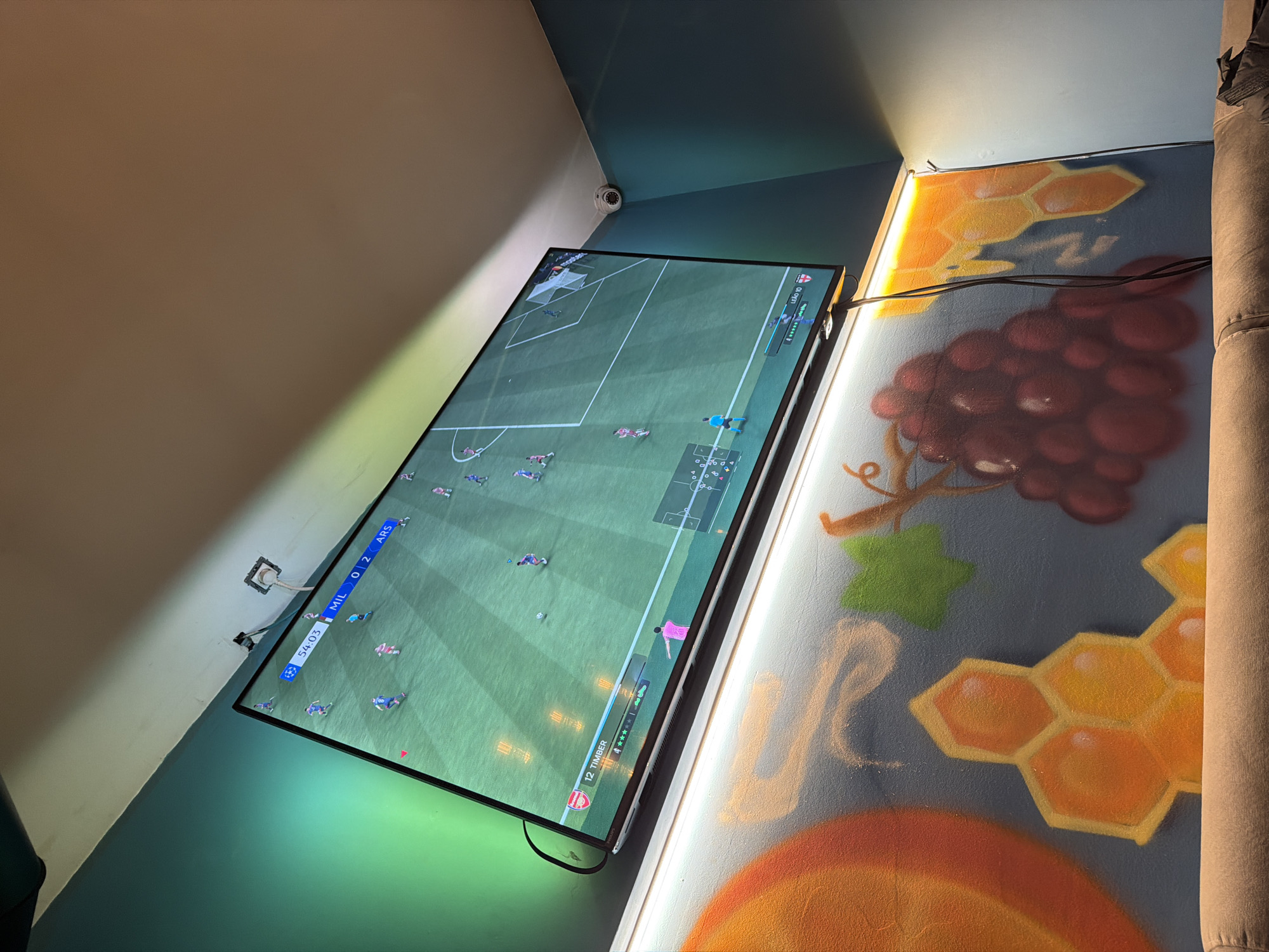 A slightly off-kilter shot shows a large flat-screen television mounted on a wall. The television is displaying a soccer game, with players visible on a green field. The score at the top left of the screen shows "MIL 0 - 2 ARS."
The wall behind the television is painted with a mural. The mural depicts a variety of food items, including grapes, honeycomb, and what appears to be a setting sun at the bottom. The color palette consists of oranges, yellows, greens, browns, and shades of grey.
To the right of the television is a neutral-colored vertical structure. A security camera is mounted on the wall near the top of the television. Electrical wires are visible running from the television along the wall.
The location is not identifiable within Sofia, Bulgaria.