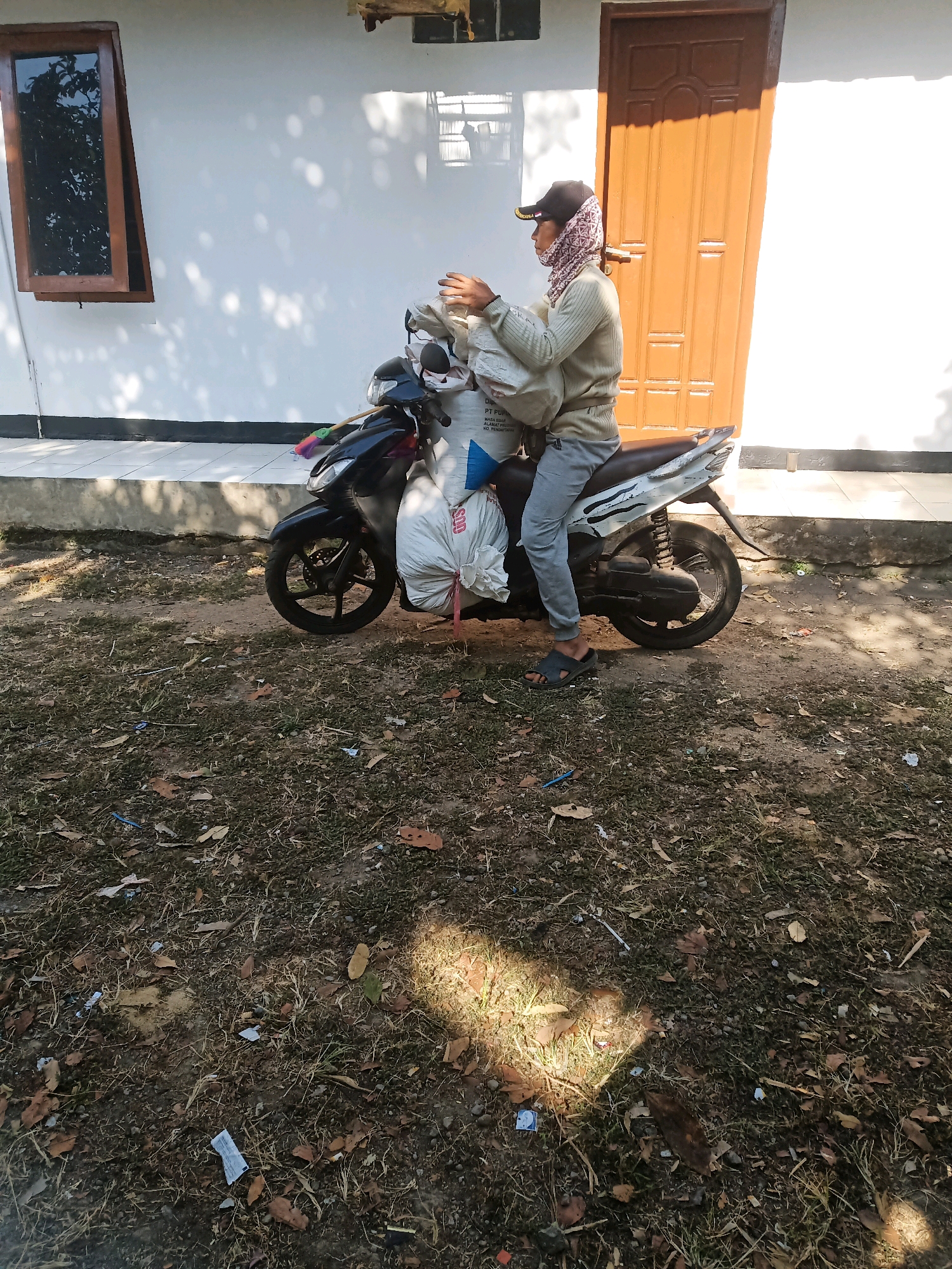 A person wearing a cap and a patterned headscarf, a light-colored long-sleeved shirt, and grey pants sits on a white and black scooter. The person is holding a white sack in their hands. The scooter is parked on a dirt and grass ground in front of a white building with a brown door and a wooden-framed window. Shadows from trees are cast on the building and the ground. The environment appears to be outdoor, possibly residential or a small business area. The scene does not provide explicit indicators of the city of Garut, Indonesia, beyond what was provided in the prompt.