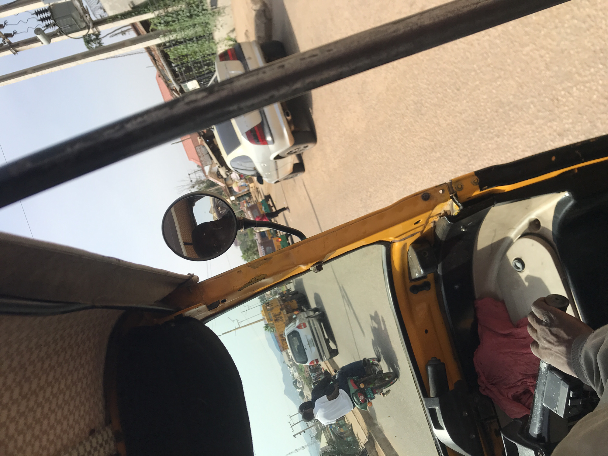 The image provides a dynamic view from inside an auto-rickshaw, looking out onto a bustling street scene during daylight hours. The vehicle's interior is visible with its yellow and black frame, a patterned seat on the left, and a round rearview mirror reflecting parts of the road and sky.
On the right side of the frame, a person's dark-skinned right hand is seen gripping a control mechanism, possibly a gear lever or throttle, which is partially covered by a crumpled red cloth. This suggests the vehicle is in motion or preparing to move, and the hand belongs to the driver.
Outside, the street is paved with a light-colored surface under a clear, bright sky, indicating sunny weather. Directly ahead and slightly to the right, a white car with a dark rear bumper is visible. The auto-rickshaw's windshield and side mirror reflections offer a wider perspective of the road, showing another white car and a person riding a green and black motorbike. The motorbike rider is wearing a white shirt and a dark helmet, positioned behind the auto-rickshaw in the reflection. In the background, buildings with reddish roofs and utility poles with power lines line the street, characteristic of an urban or semi-urban environment. No specific text is clearly legible in the image. The scene captures a typical moment of travel in Jos, Nigeria.