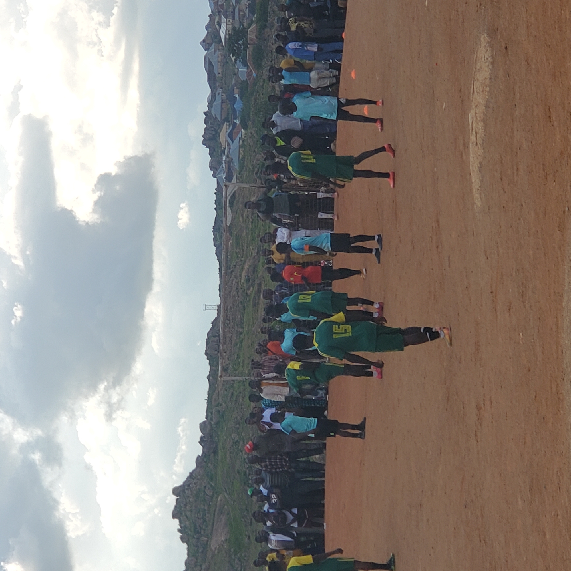 This image depicts an outdoor football match taking place on a dirt field in Jos, Nigeria. A group of young men, dressed in green and yellow football kits, are on the field, some actively playing and others observing. A crowd of spectators, varying in age and attire, are gathered around the perimeter of the field, watching the game. In the background, a makeshift goalpost with a net is visible. The landscape behind the field is characterized by rocky hills dotted with residential buildings, suggesting an urban or semi-urban setting within Jos. The sky is overcast with dramatic clouds, casting a diffused light on the scene. The ground appears dry and dusty, typical of a football pitch in a region with limited vegetation.