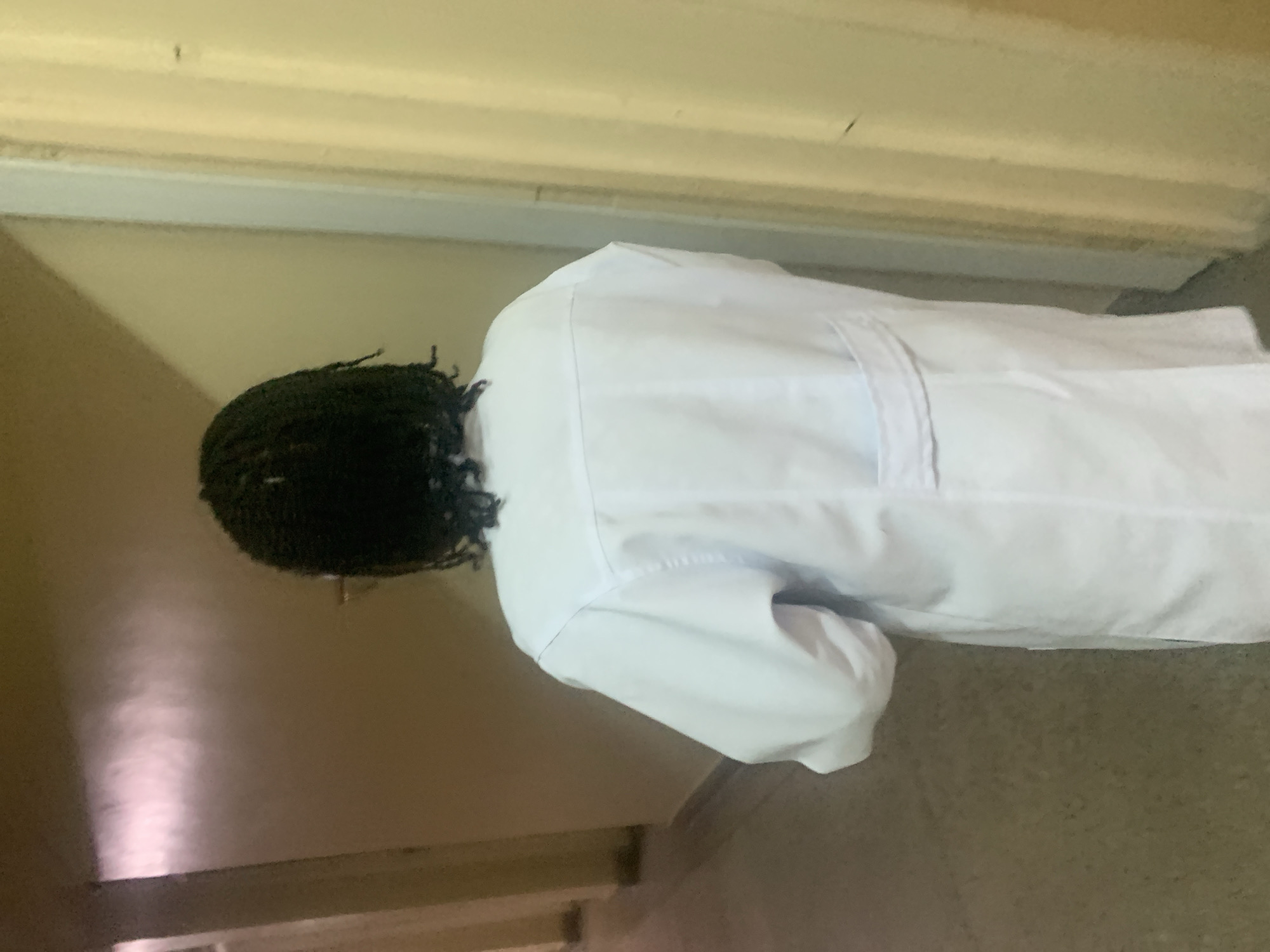 A person with dark, braided hair is wearing a white lab coat in a hallway. The walls are painted a light yellow color with a horizontal strip near the top. The location is likely inside a building, possibly a hospital or clinic. The city of Dar es-Salaam, Tanzania, cannot be confirmed from the image, though it is stated in the prompt.