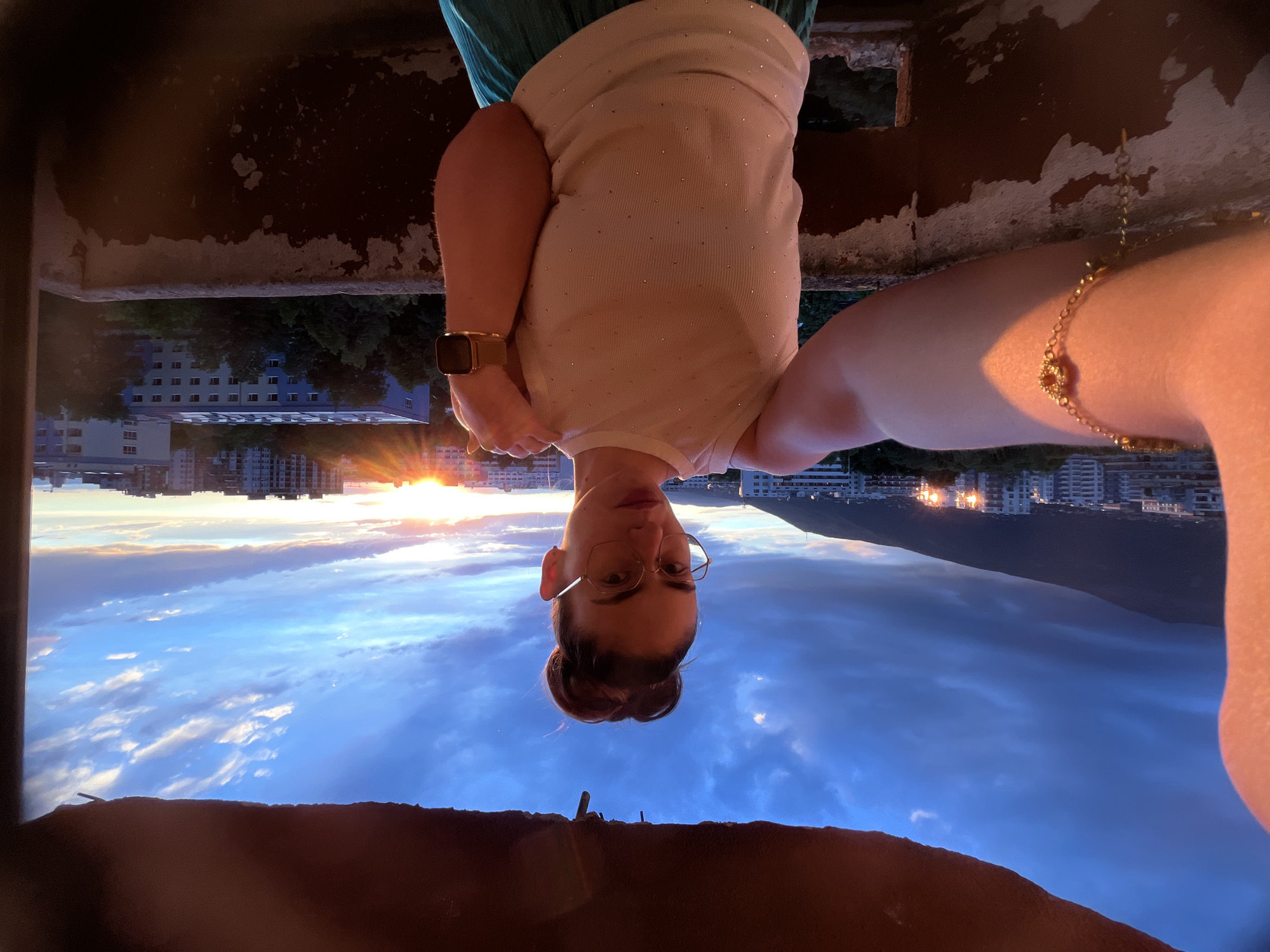 A woman is taking a selfie on a balcony with a cityscape and sunset in the background. The woman, with her hair in a bun and wearing glasses, is dressed in a white, sleeveless top and a turquoise skirt. She has a gold bracelet on her left wrist and a smartwatch on her right wrist. The balcony appears weathered and aged. The sky is a mix of blues and oranges, with clouds, suggesting it is either sunrise or sunset. The cityscape consists of buildings and a mountain range in the distance.