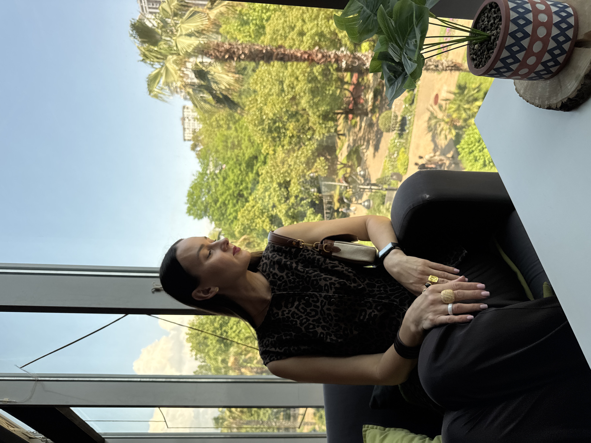 A woman with dark hair is seated in a dark armchair, looking out a large window. She is wearing a sleeveless, leopard-print top and dark pants. She has several rings on her fingers and a watch on her left wrist. A handbag with a tan strap is slung over her right shoulder. Through the window, a lush green park with palm trees and buildings can be seen under a clear blue sky. The scene is illuminated by natural light. The presence of palm trees and buildings in the background suggests an urban park setting within Sochi, Russia. A potted plant with large green leaves sits on a white table in the foreground.