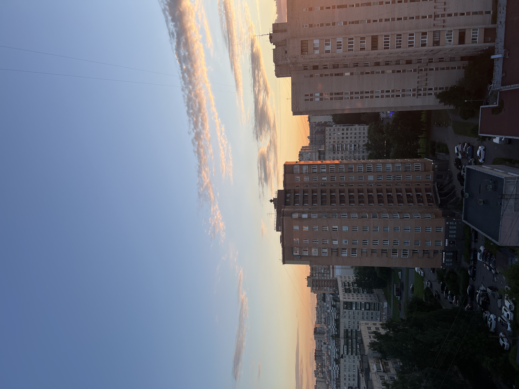 The image captures a city skyline at dusk in Unknown, Russia. The sky is a gradient of blue, transitioning to a soft orange and pink hue near the horizon, with scattered clouds catching the last light of the sun. Dominating the foreground are several tall, brick residential buildings, typical of Soviet-era architecture. One building, prominently featured on the left, is light brown with a regular pattern of white-framed windows. To its right, another taller building, constructed of lighter colored bricks, has a similar window arrangement and is illuminated by the setting sun, casting a warm glow on its facade. In the distance, a densely packed cityscape of more residential blocks stretches across the horizon, interspersed with trees and greenery. The lower portion of the image shows a street level view with parked cars, a road, and more apartment buildings, some with visible signage. The scene is quiet and still, depicting a common urban environment during the evening hours.