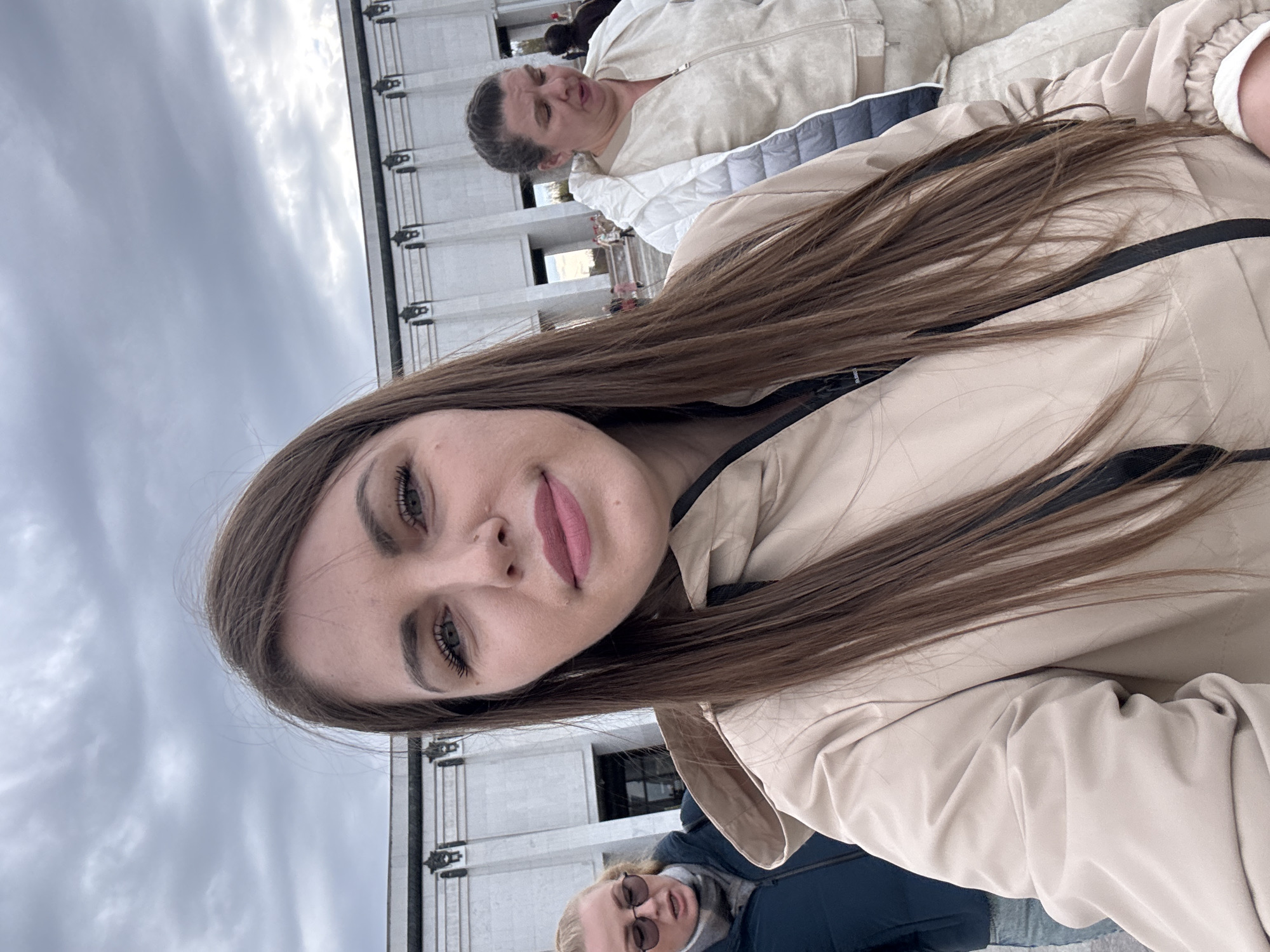 The image shows a selfie of a woman with long brown hair wearing a beige jacket. Behind her are two other people and a building with multiple windows and decorative metalwork. The cloudy sky suggests an overcast day. Based on the architectural style of the building, it is likely taken in Moscow, Russia.