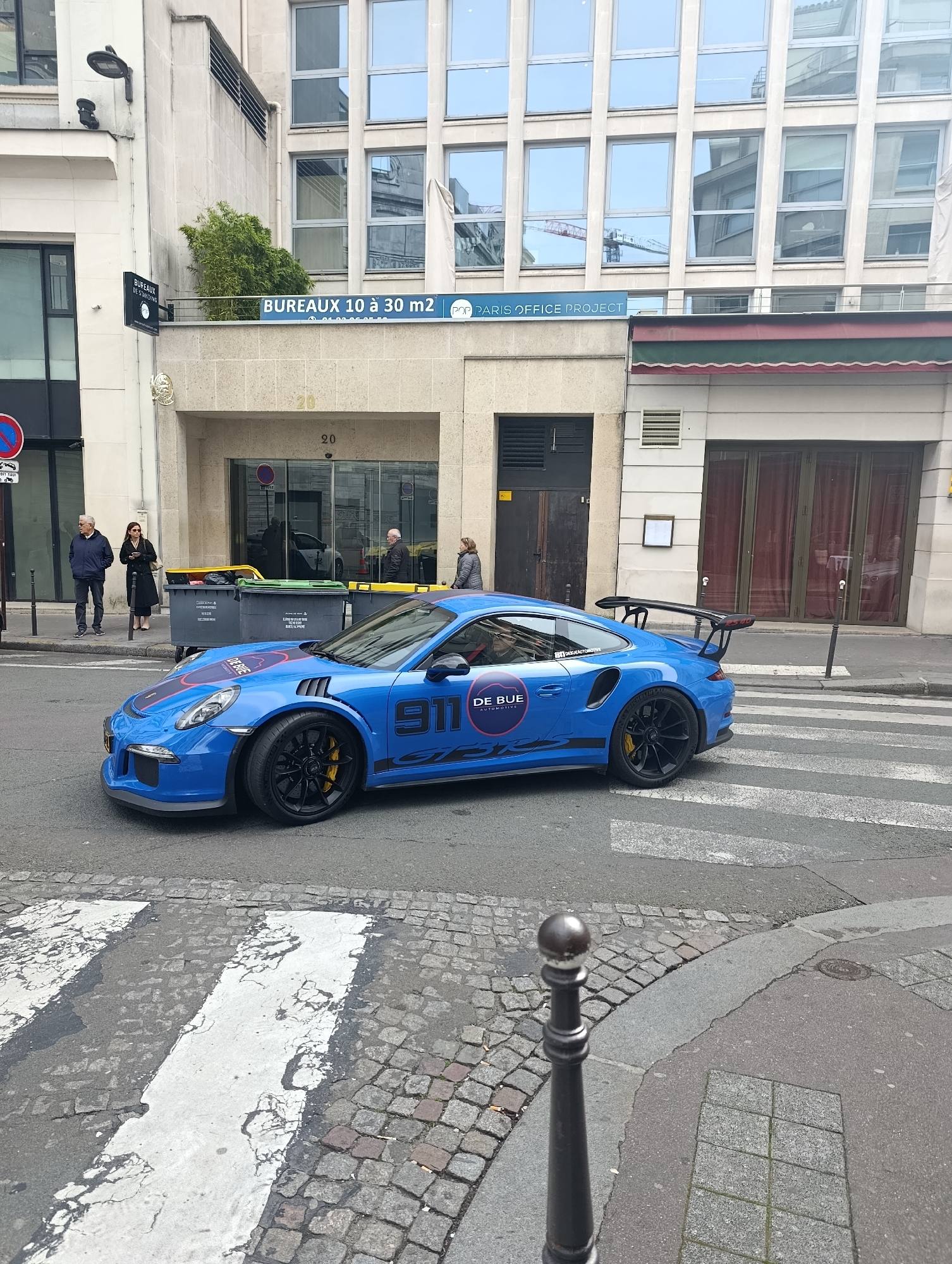 The image shows a blue Porsche 911 GT3 RS car parked on a zebra crossing in Paris, France. The car features the "DE BUE" logo and the number "911" prominently displayed. The car is positioned on a cobblestone street corner, with a black bollard visible in the foreground.
In the background, several people are seen on the sidewalk near a building complex. The building has the signage "BUREAUX 10 à 30 m2" and "POP PARIS OFFICE PROJECT" visible above its entrance, and there is a sign "BUREAUX DE STANDING" to the left. There are some large waste containers near the curb. The building's facade includes multiple windows, some reflecting the surrounding environment.