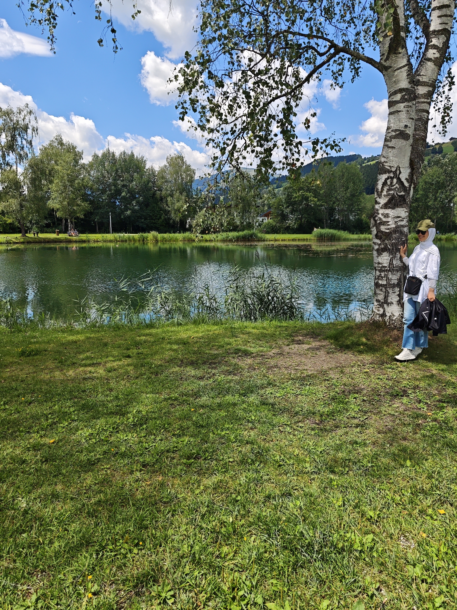 The image shows a woman standing next to a birch tree by a lake in Mittersill, Austria. The woman is wearing a white shirt, blue jeans, a green cap, and white sneakers. She is holding a black bag in her right hand and a jacket draped over her arm. She has a sling bag across her chest. The lake is calm and reflects the blue sky and white clouds. Trees and hills are visible in the background. The foreground is a grassy lawn with some shadows. The overall scene depicts a peaceful outdoor setting.