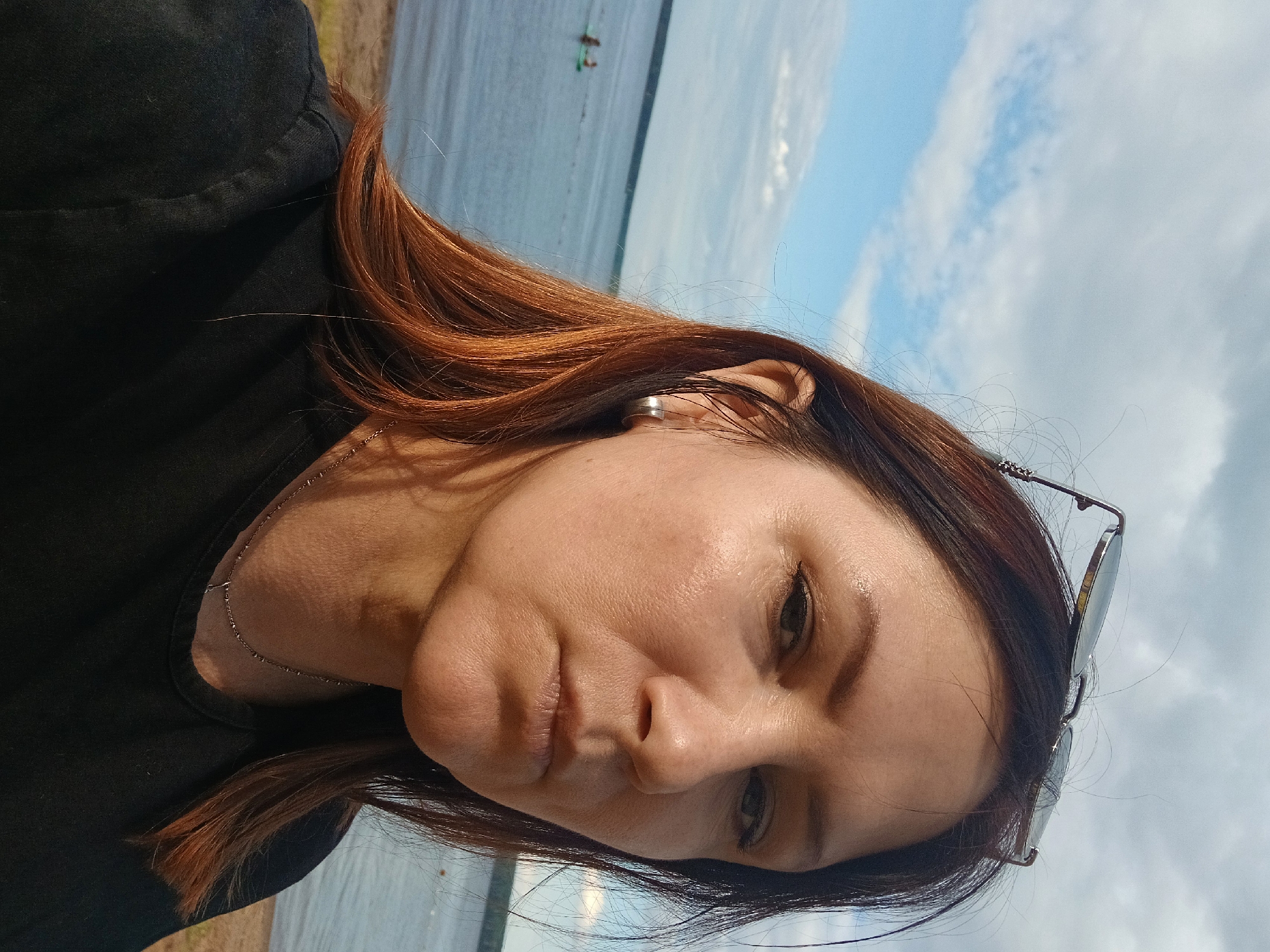 A medium shot shows a woman with shoulder-length auburn hair and sunglasses perched on her head. She is looking directly at the camera with a neutral expression. She is wearing a black t-shirt and a silver necklace. Behind her, a body of water is visible, with a sandy shore in the foreground and a few people in the water in the background. The sky is cloudy with patches of blue. The setting is likely the Kama River in Chaikovsky, Russia, given the presence of a large body of water and the context of the city.