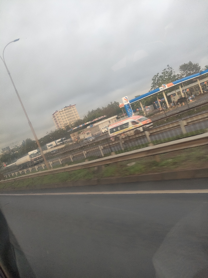 The image depicts an outdoor urban scene under an overcast sky, captured from a moving vehicle. In the immediate foreground, a dark asphalt road surface is visible, featuring a faint yellow lane demarcation line. A metal guardrail runs horizontally across the mid-ground, separating the foreground road from an elevated multi-lane highway or railway line.
On the elevated thoroughfare, vehicles are in transit, exhibiting motion blur. A white and orange passenger vehicle, possibly a train or large bus, moves from right to left. A white tanker truck is also visible, along with several smaller cars and vans. A tall, dark lamppost stands on the far left.
Beyond the elevated roadway, a busy OLA Energy gas station is prominent. It features a blue and white canopy sheltering multiple fuel pumps. Several individuals are present at the gas station, some near the pumps and others around an adjacent building structure, indicating active patronage. Behind the gas station and to its left, a multi-story, light-colored building with numerous windows rises against the horizon. Various trees and green vegetation are scattered throughout the scene. The setting is identified as Theta ward, Kenya.