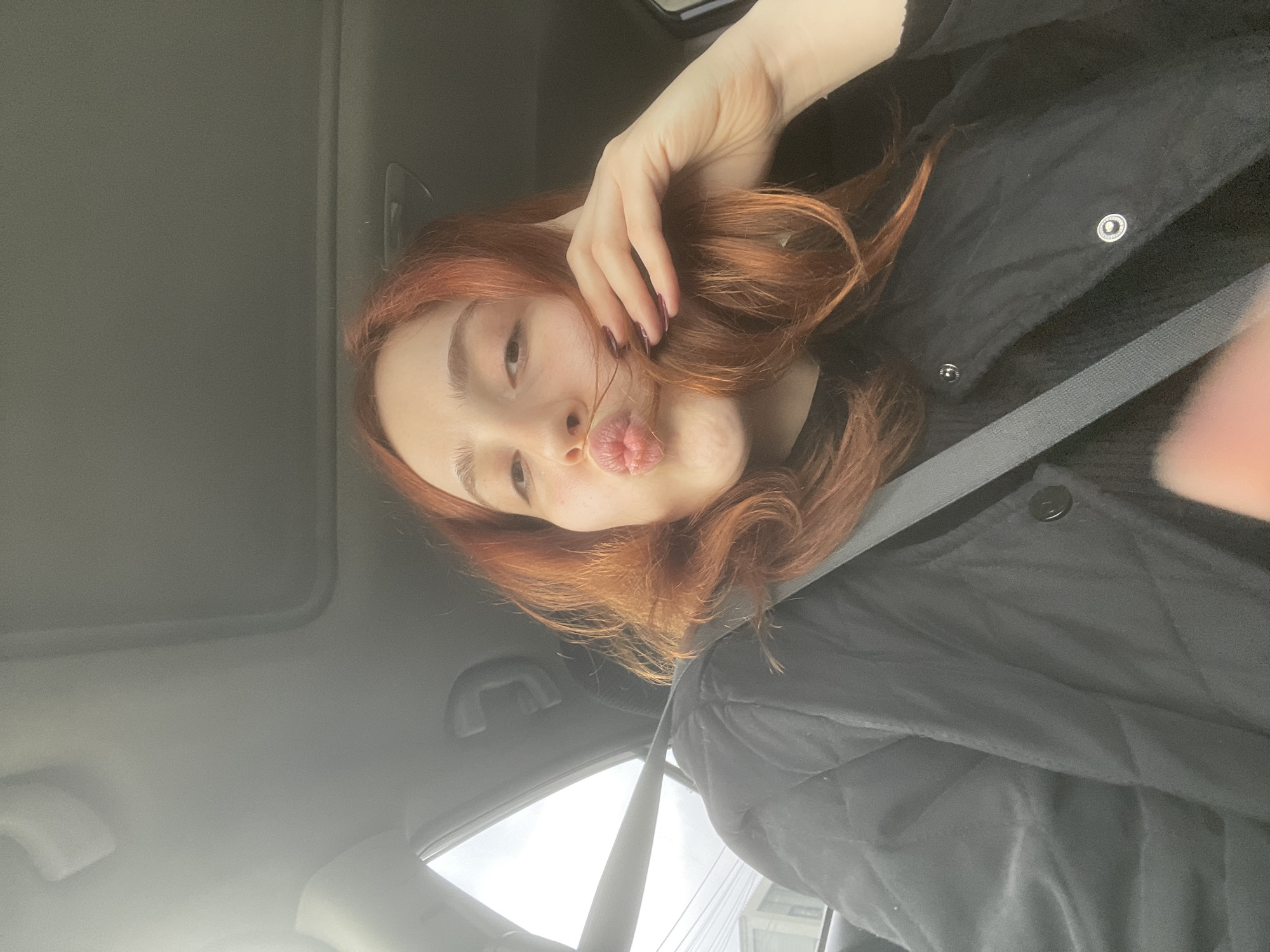 A young woman with red hair poses in a car, wearing a black jacket and a seatbelt. She is making a kissing face and has her hand in her hair. The car appears to be in motion, with a glimpse of buildings and sky visible through the windshield. The interior of the car includes a sunroof and standard car fittings. The location cannot be determined to be Tbilisi, Georgia based on visual information.