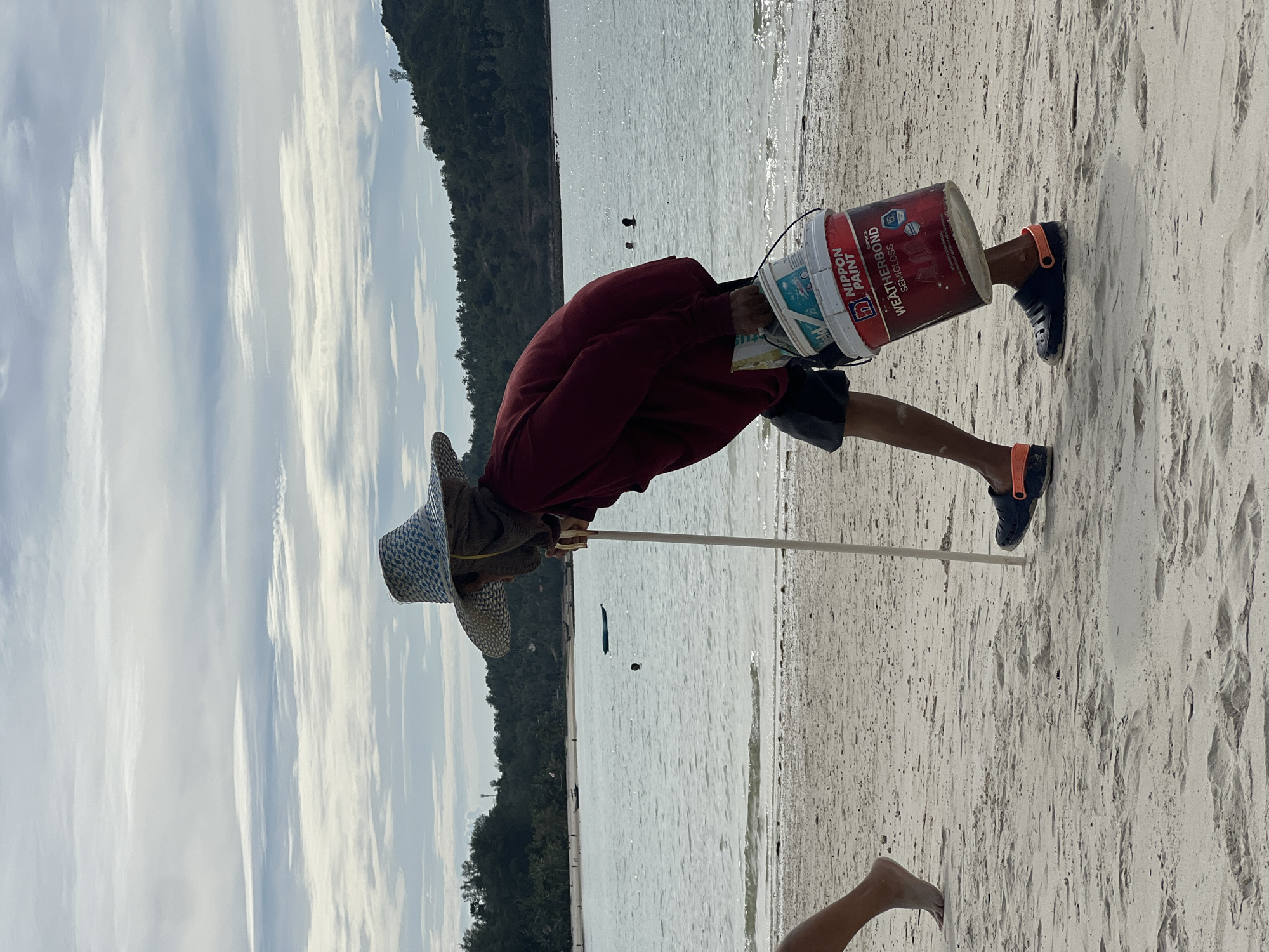 A person wearing a wide-brimmed hat and a maroon shirt walks along a sandy beach in ตำบลเกาะพะงัน, Thailand. They are carrying two buckets, one red and one white, by a handle. The person is also using a long, thin pole as a walking aid, holding it with their right hand. The other hand is holding the buckets. The beach is wet near the water's edge, and the ocean is visible in the background. There are trees and hills on the far side of the water. Another person's legs are visible in the foreground on the left, walking in the same direction as the main subject. The sky is overcast with visible cloud formations.