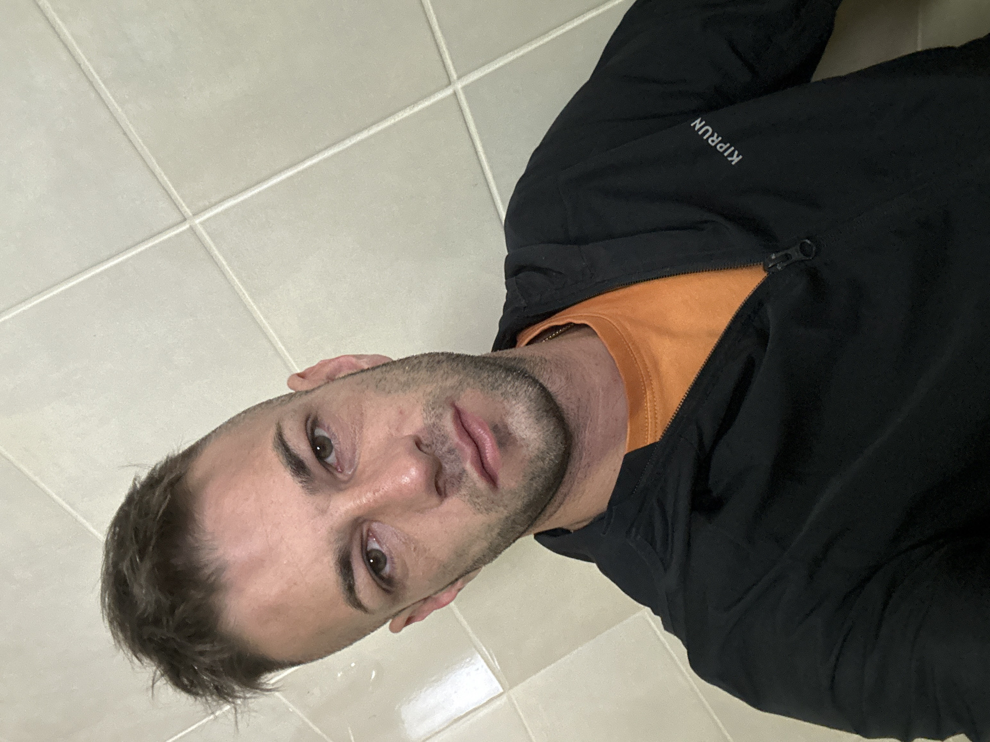 The image is a close-up selfie of a man with short, dark hair styled with a faded haircut, and stubble on his face. He is wearing an orange t-shirt and a black zipped jacket with the brand "KIPRUN" visible on the left side of the chest. The background consists of light-colored square tiles with grout lines. The man's expression is neutral. The scene does not provide any information or context that could place it in Piperevo, Bulgaria, or any other specific city.