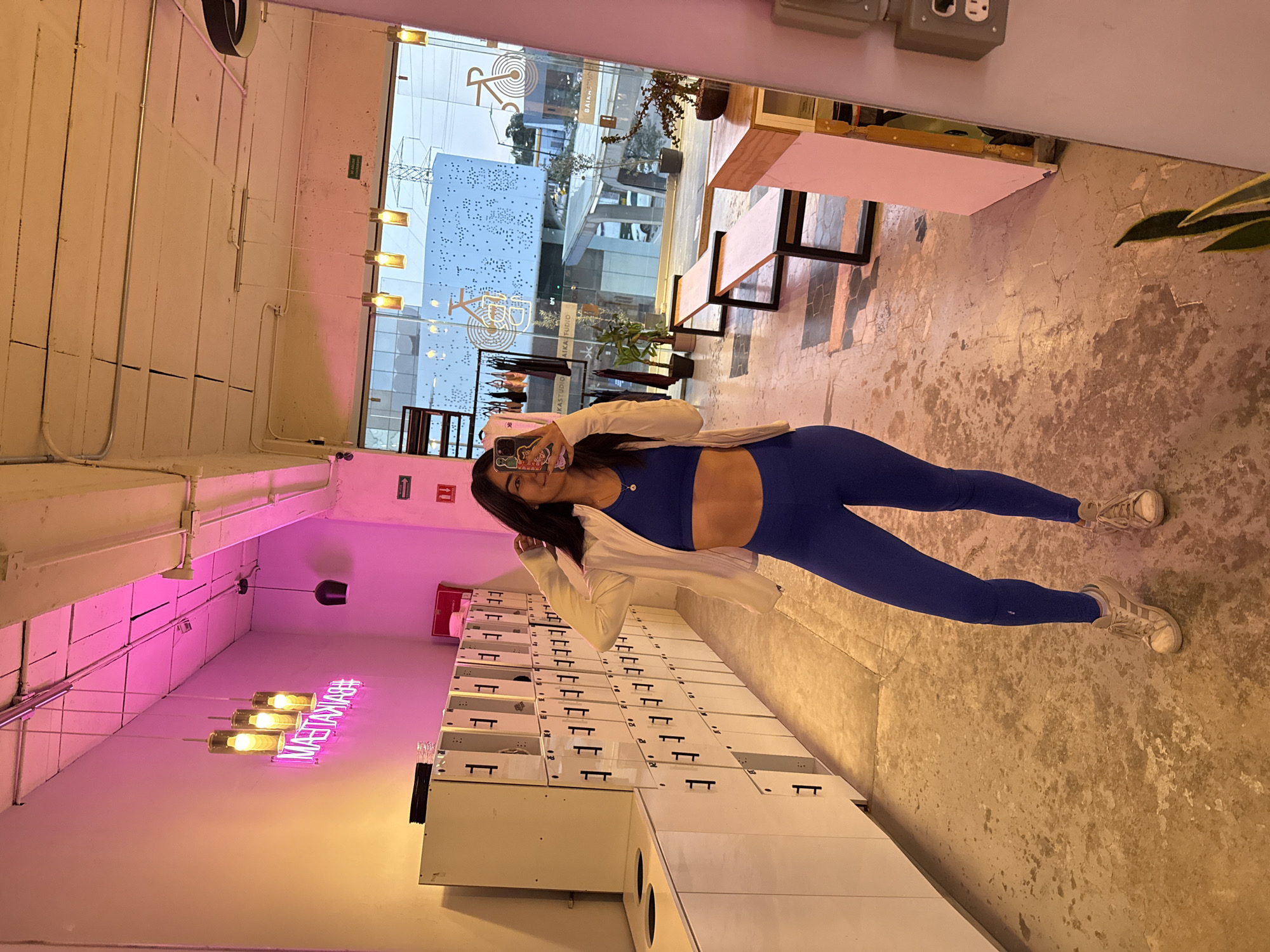 A young woman wearing a blue workout set and a light-colored jacket stands in a locker room with white lockers and a pink neon sign. She is taking a selfie in a mirror that reflects a view of the street outside, showing a building with a logo that reads "IKASTUDIO IKAFUDIO BAIKA." The location is likely in Puebla, Mexico. The room has a modern aesthetic with concrete floors and exposed ductwork.