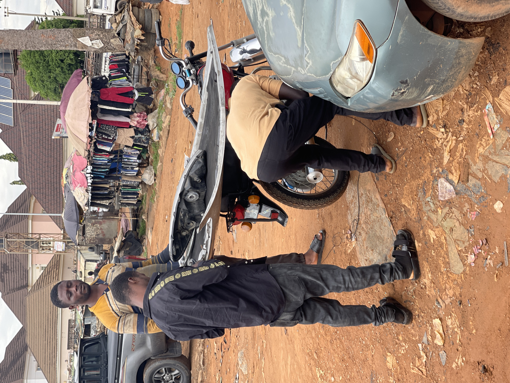In Karshi, Nigeria, two men are working on a motorcycle and a damaged blue car. One man, wearing a black jacket with a distinctive pattern on the shoulders, stands facing the camera. A second man, wearing a tan shirt and black pants, is bent over, apparently working on the motorcycle or the car. The front of the motorcycle has been removed and is leaning against the bike. The car's front bumper is damaged. They are on a dirt road with scattered debris. In the background, there is a display of clothing under an umbrella, a parked gray pickup truck, and various buildings.
