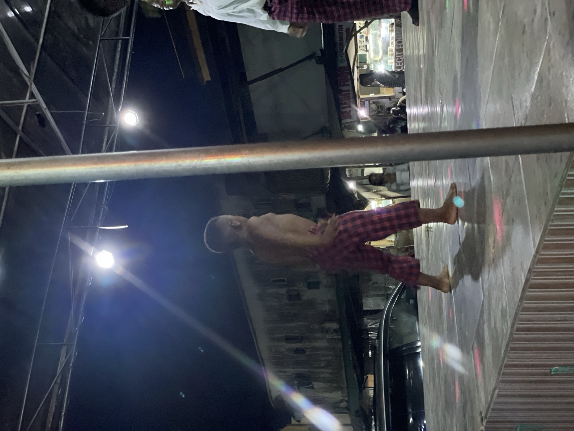 A street scene in Ado, Nigeria, presents a partially nude man wearing red plaid shorts, standing barefoot on a concrete sidewalk adjacent to a closed storefront with a corrugated metal roll-down gate. He appears to be performing or posing. The street is illuminated by overhead lights, including one mounted on scaffolding to the left, casting strong directional light. Other people are visible in the background near storefronts further down the street. A car is parked beside the sidewalk.
