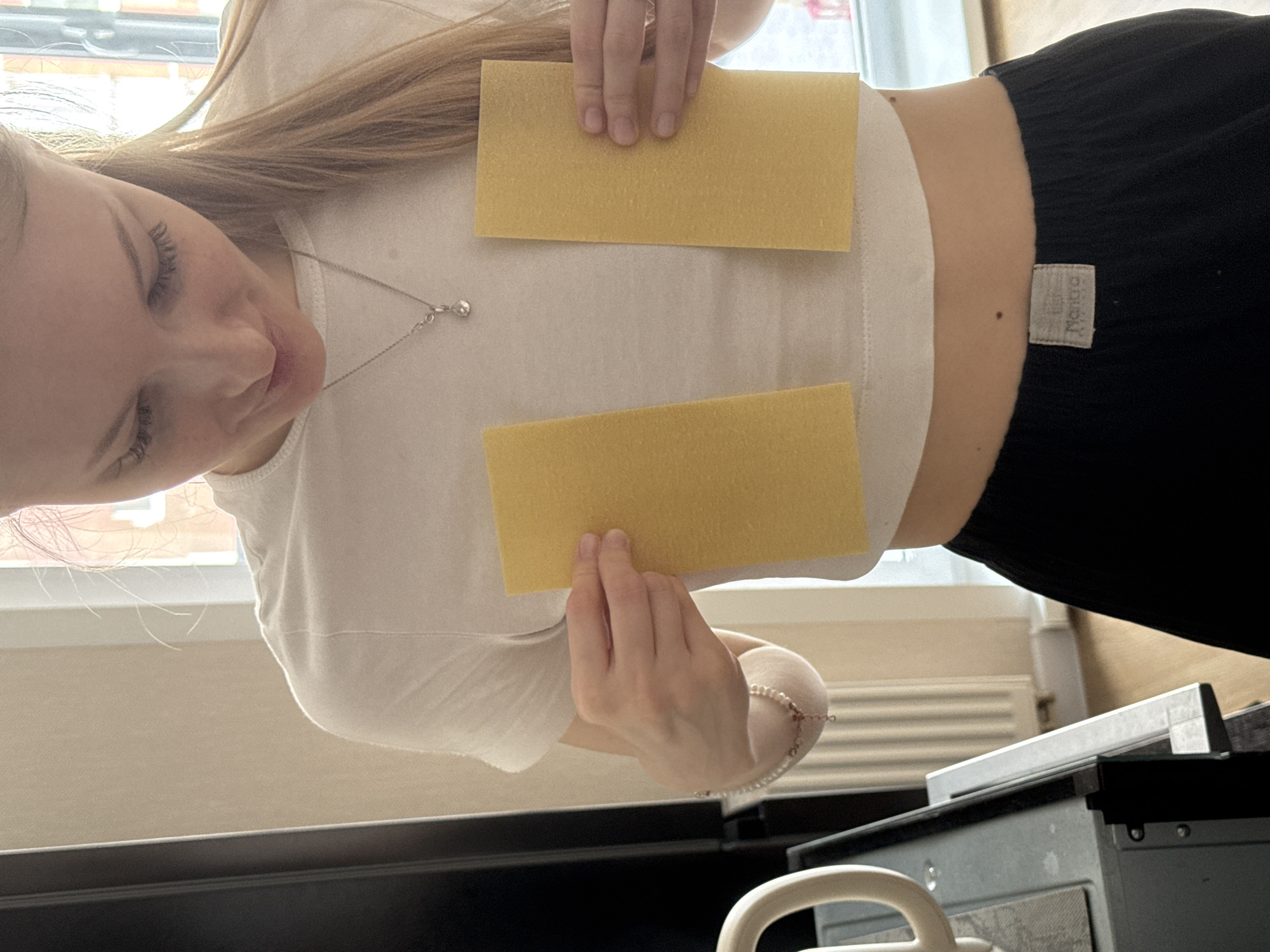 A young woman is depicted indoors, holding two rectangular, uncooked lasagna sheets. The image is oriented such that her head is on the left, looking down towards the two yellow sheets held vertically against her white t-shirt. Her long, light brown hair flows across her back and shoulders. She is wearing a white t-shirt, black pants, a silver chain necklace with a small clear pendant, and a light-colored beaded bracelet on her right wrist.
The setting appears to be a brightly lit room, likely a kitchen or dining area, with strong natural light coming from a window behind her. Through the window, parts of a reddish-brick building can be seen. A white radiator is visible beneath the window. In the foreground, a dark surface, possibly a kitchen counter or appliance like an oven, with a metallic handle, is partially visible.
The woman has a neutral expression, with visible eyelashes. A small, dark mole is located on her right side, near her waist. A small, light-colored fabric tag is visible on the waistband of her black pants, with text that appears to be "Melange." The bright light suggests it is daytime. The scene is likely taking place in a domestic environment in Perm, Russia.