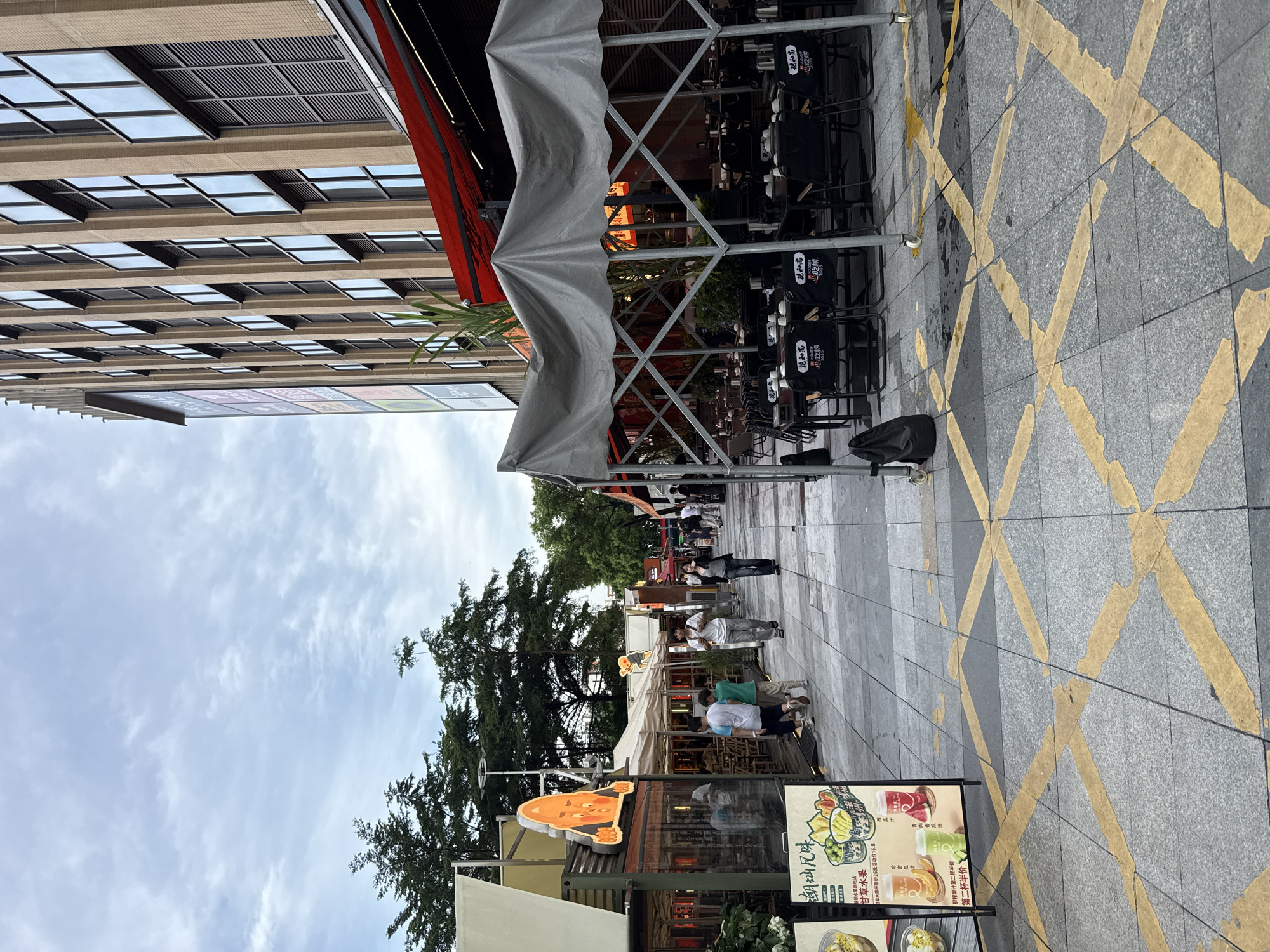 In the city of Haizhu District, China, an outdoor dining area is depicted under a cloudy sky. The scene features a paved walkway with yellow crosswalk markings. On the right, a modern building with numerous windows rises, partially obscured by a red awning. In front of the building, several tables and chairs are set up under a large, grey tent. To the left, a row of food stalls and small restaurants line the walkway, some with outdoor seating and awnings. A sign displaying images of fruit and text in Chinese is visible in the foreground. Several people are walking along the walkway, some individually and some in small groups. The atmosphere appears casual and relaxed, suggesting a lively commercial or pedestrian zone.